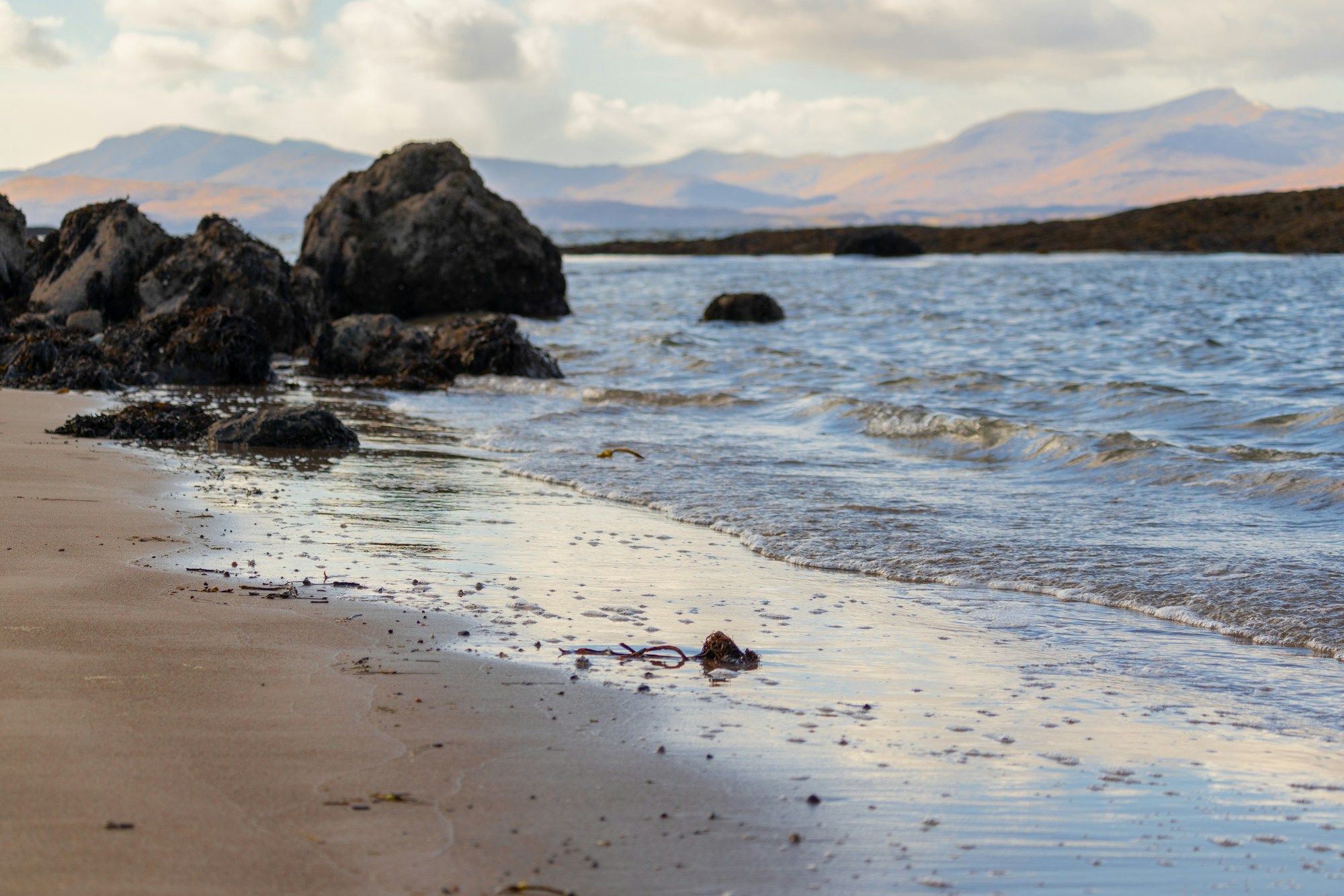 An empty rocky and sandy Scottish beach at low tide — dark seaweed-covered rocks, calm sea, and a hazy silhouette of low mountains across the water in evening light. Nobody in sight.