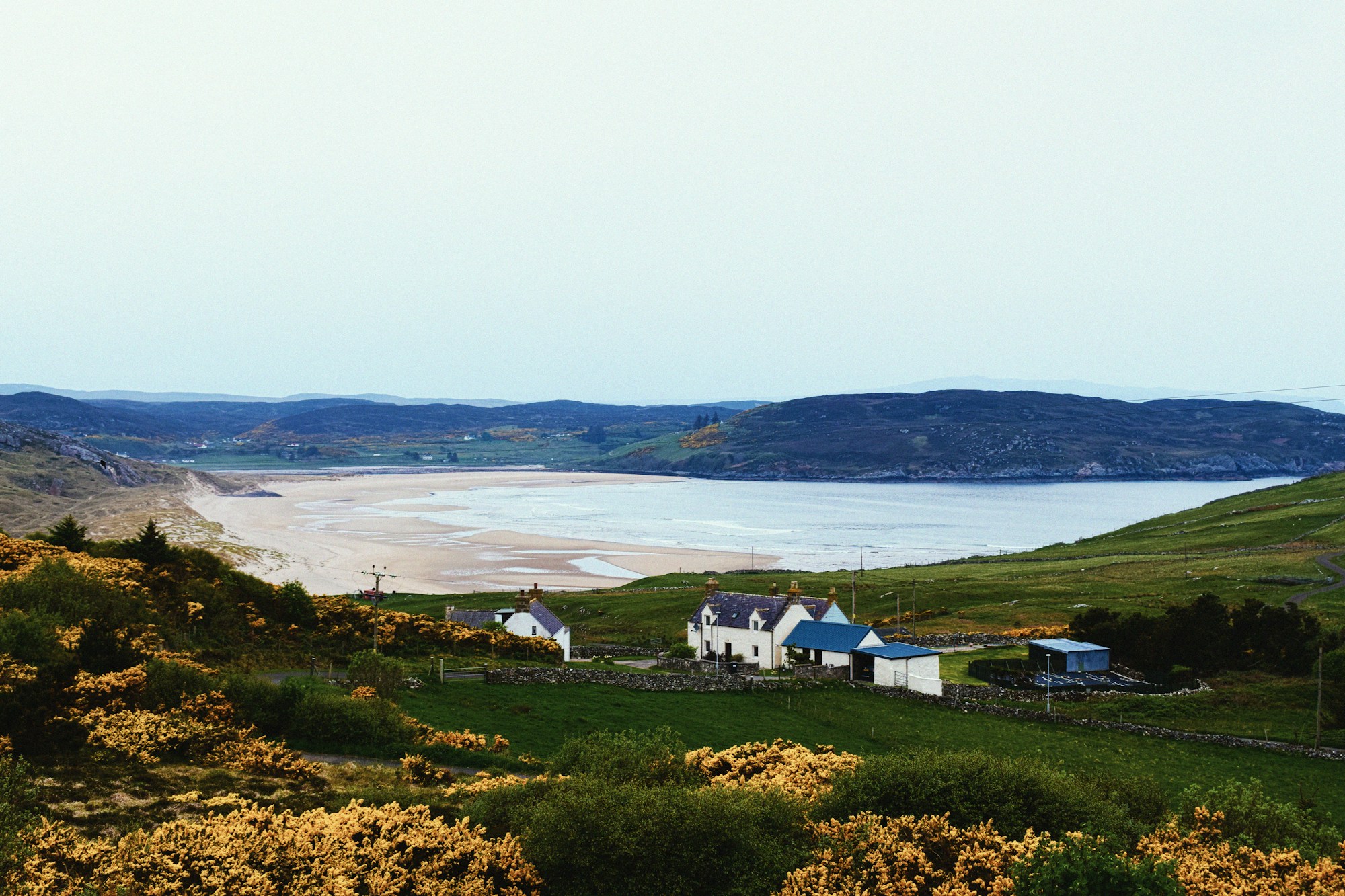 A whitewashed Highland farmhouse and its outbuildings on green pasture above a curving sandy bay, ringed by low moorland hills, yellow gorse in the foreground.