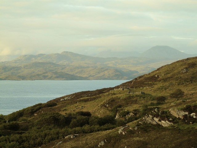A high view from the Drumbeg viewpoint over a sea loch and low hills, distant mountains hazy on the horizon.