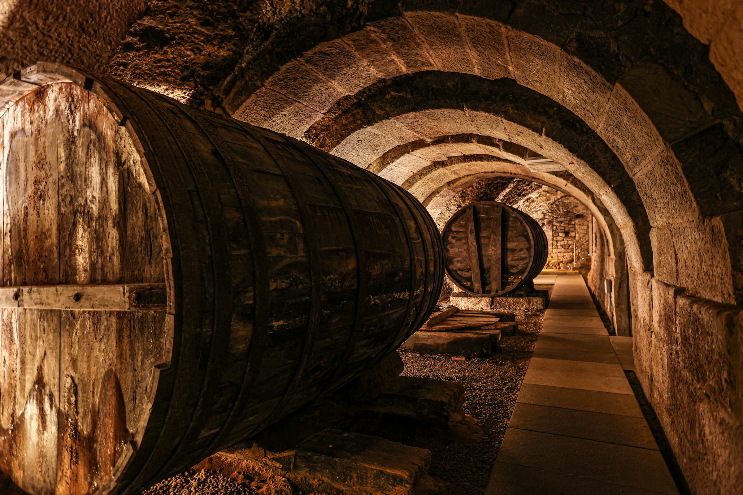Une cave à vin souterraine voûtée en pierre, de grands tonneaux de bois alignés sous un éclairage chaud.