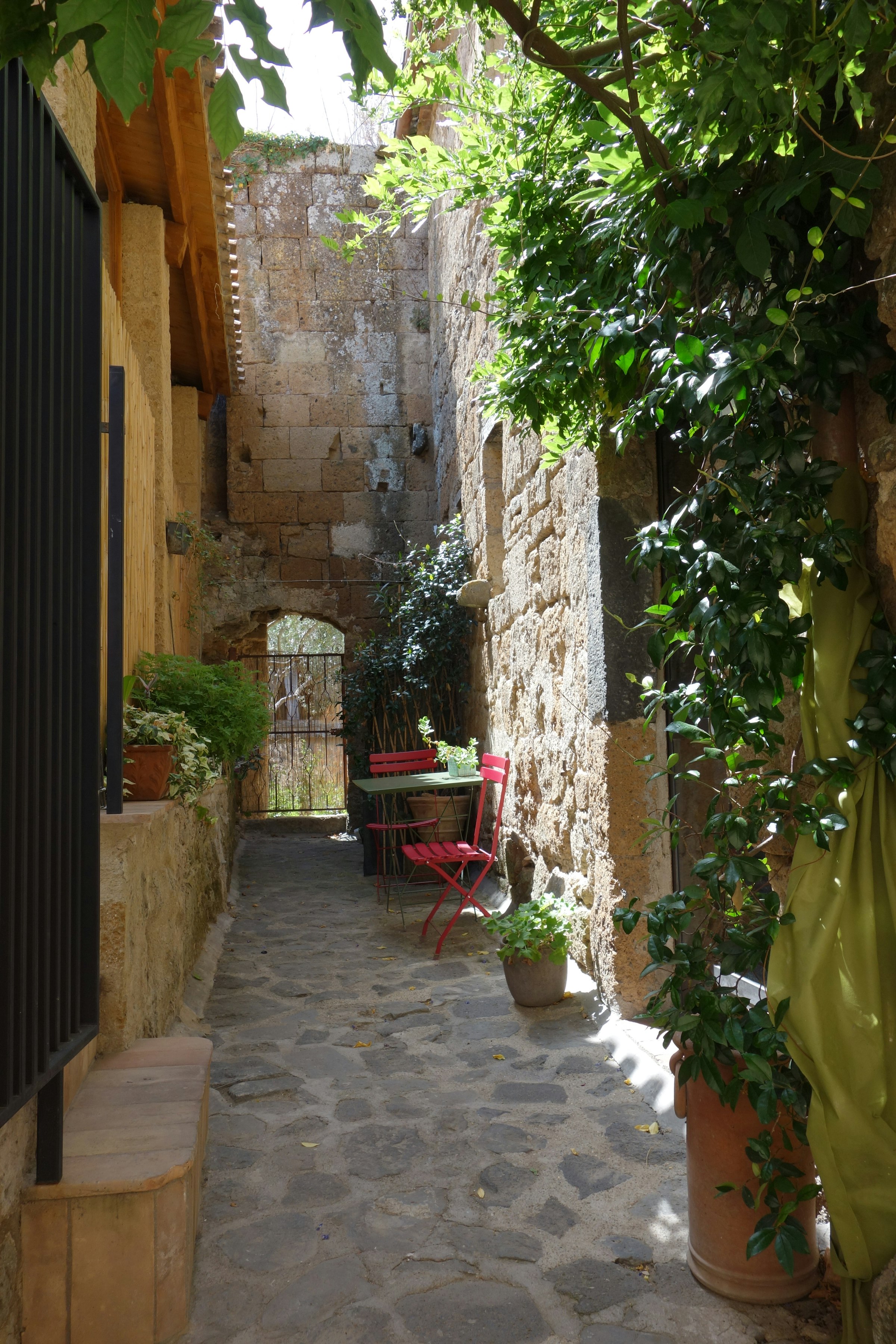 Une ruelle-cour pavée en pierre, une table et des chaises sous une voûte de verdure, des murs de pierre ancienne.