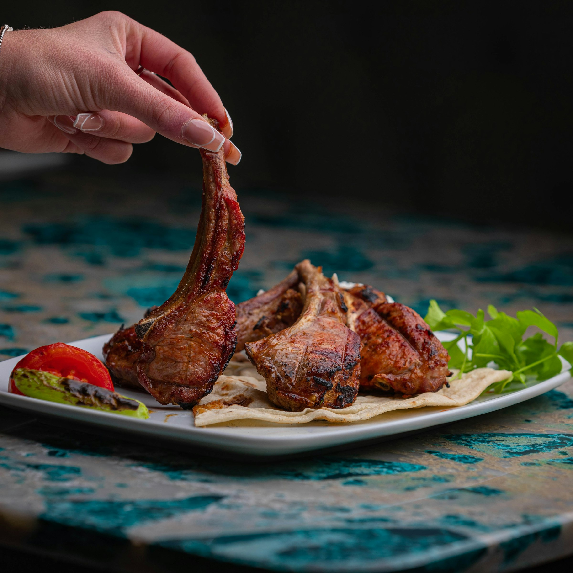 Une main soulève par l'os une côtelette d'agneau grillée (paidakia) : plusieurs côtelettes dorées et charbonnées dressées sur du pain plat, avec une tomate grillée et de la roquette