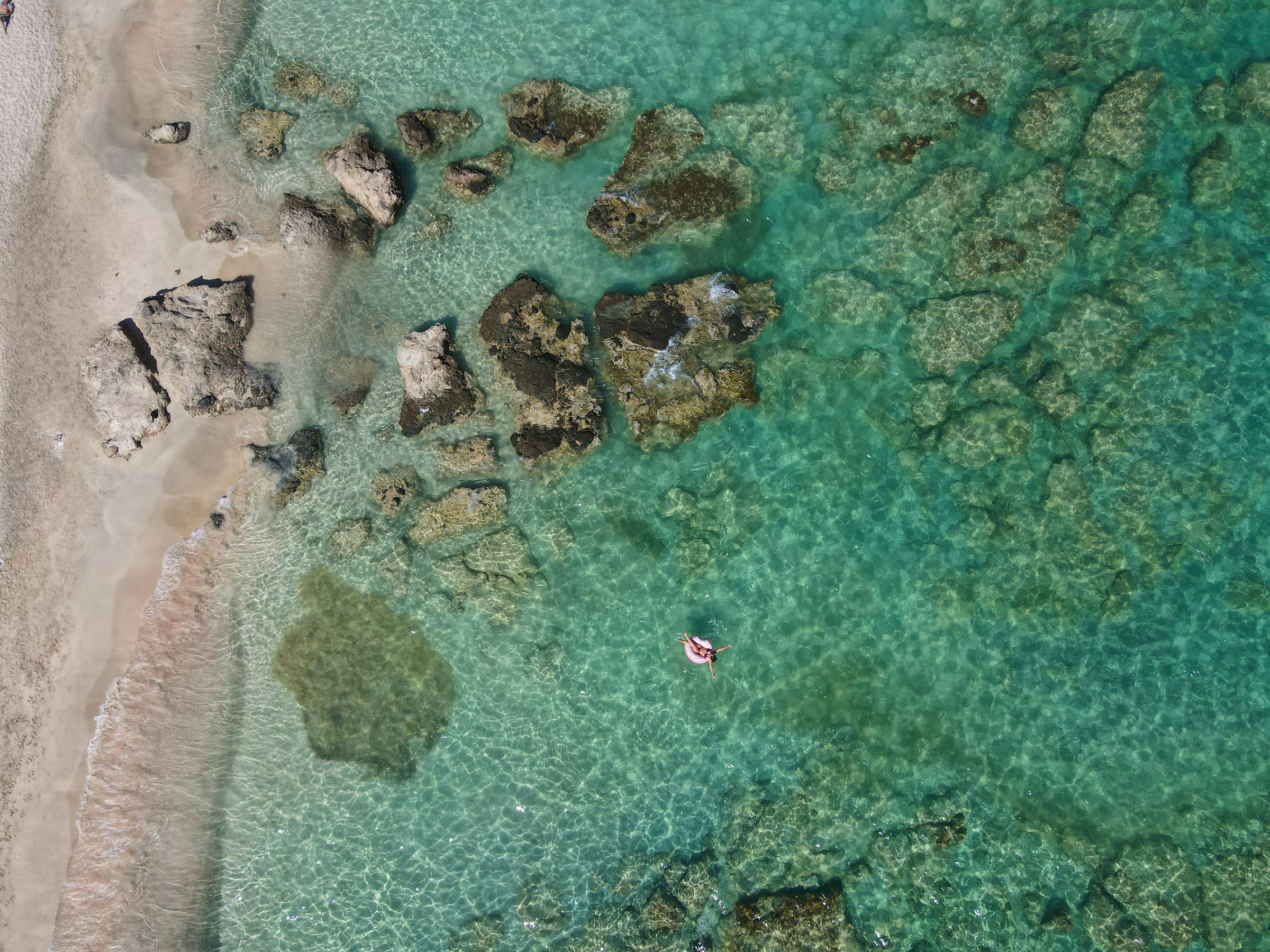 Vue aérienne d'Elafonisi, au sud-ouest de la Crète : une eau translucide vert émeraude et turquoise sur des hauts-fonds parsemés de rochers, bordée d'une plage de sable pâle