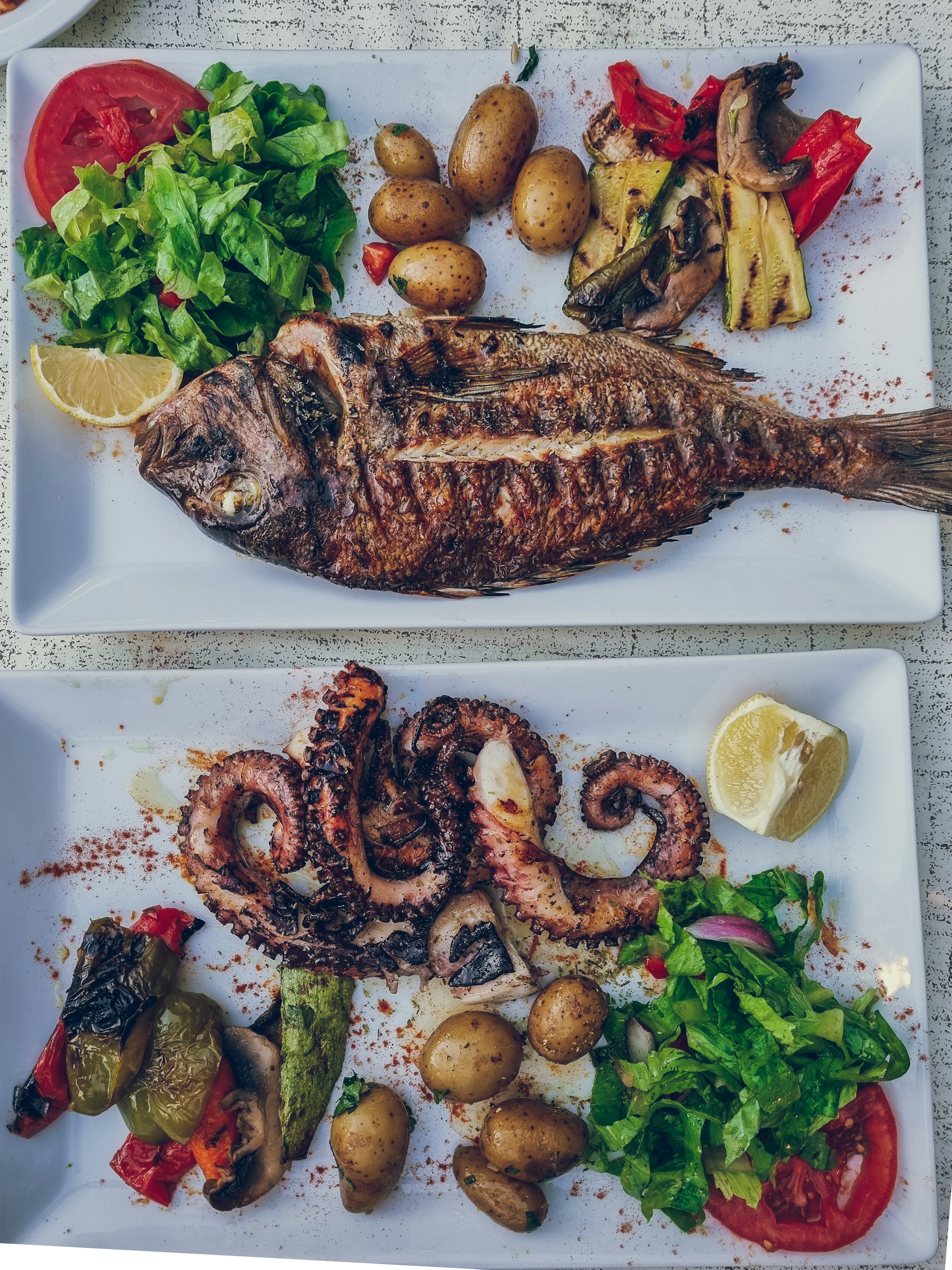 Un poisson entier grillé et un poulpe grillé au charbon, servis sur des assiettes blanches avec des quartiers de citron, dans une taverne grecque au bord de la mer