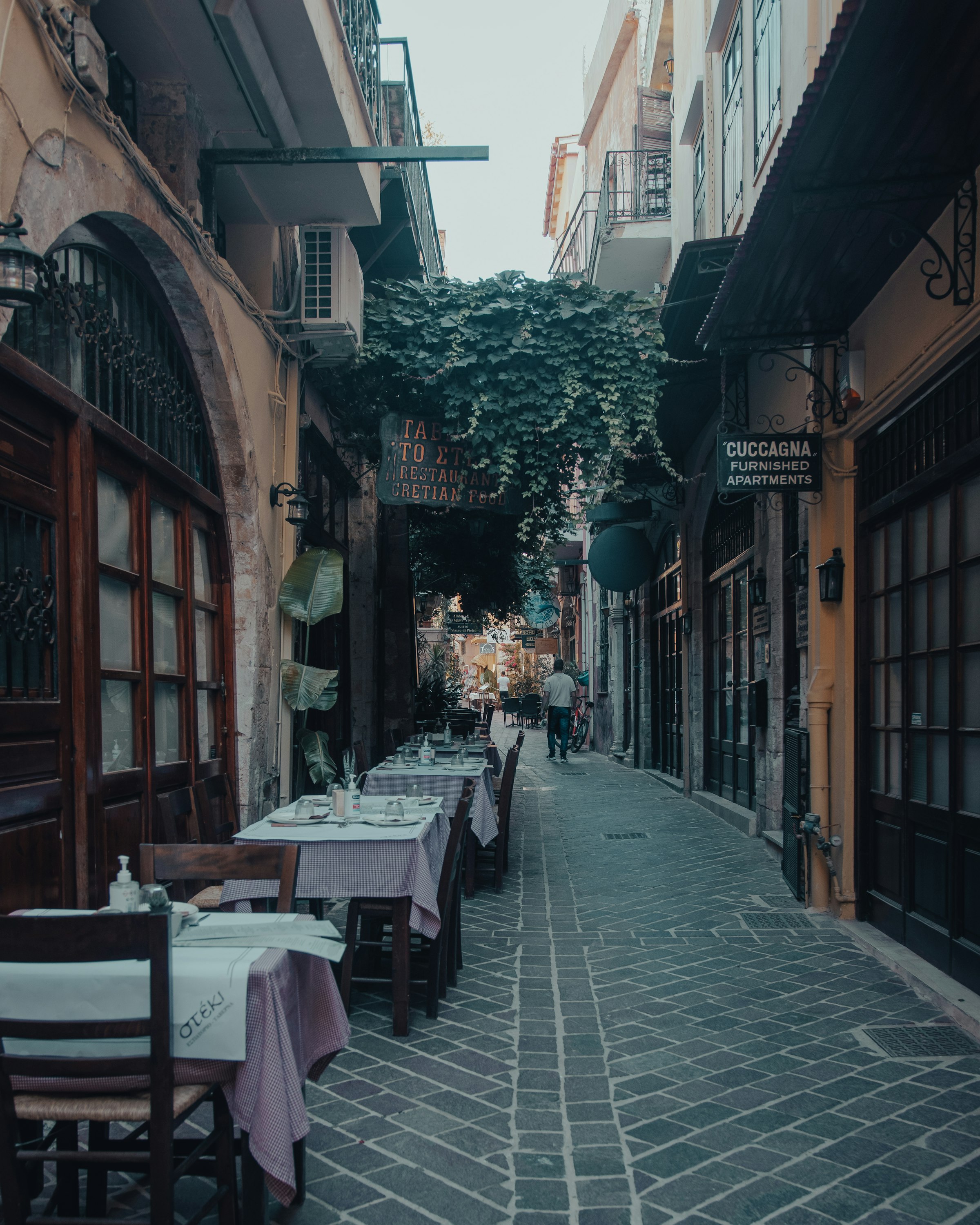 Une ruelle pavée de la vieille ville de Chania à la tombée du jour : des boutiques et tavernes aux enseignes de fer, des lanternes, et un passant au loin entre les murs anciens