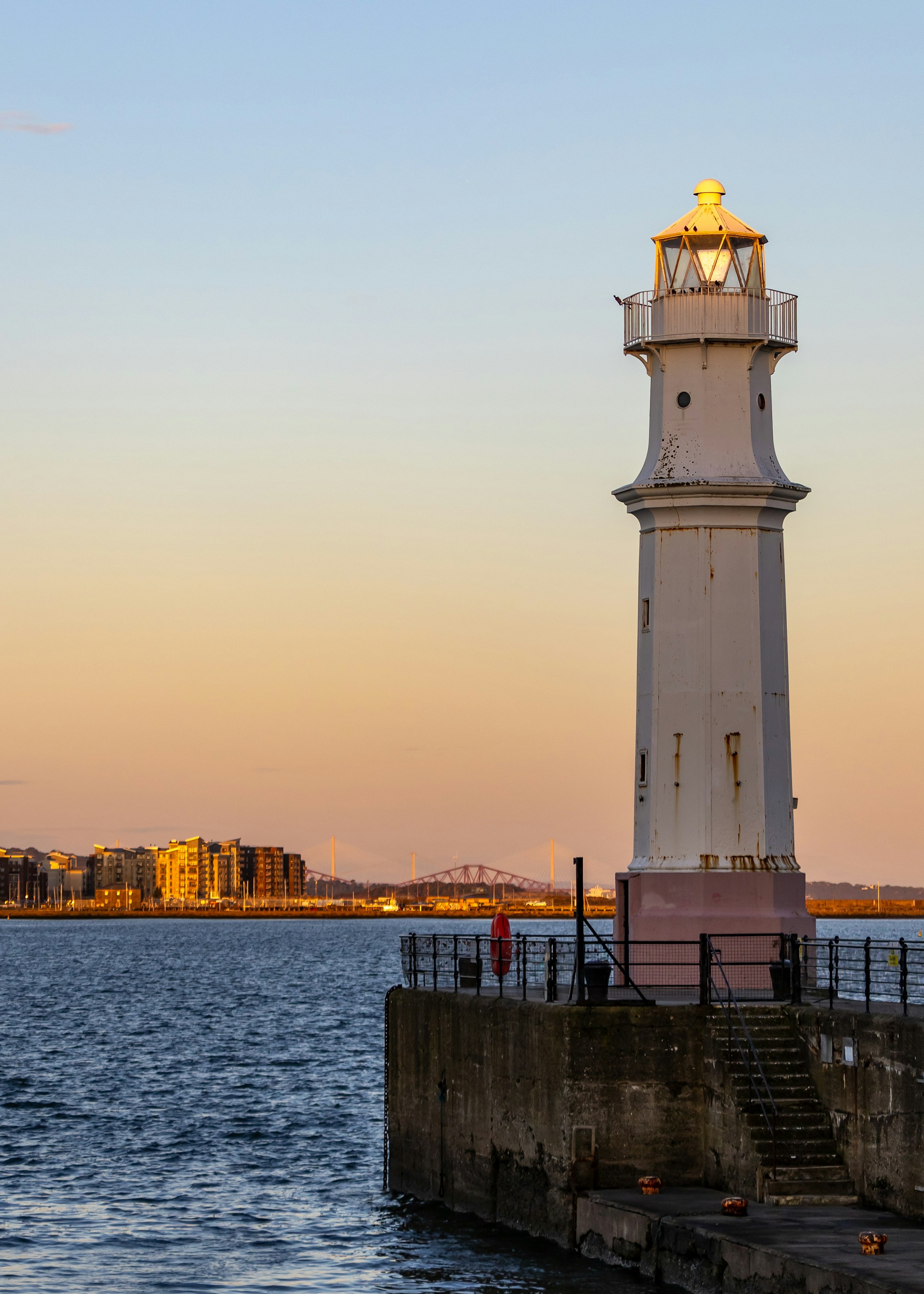 A tall white lighthouse with a rose-tinted stone base and a yellow lantern room at the end of a stone harbour pier at sunset, calm estuary water beyond, with waterside apartments and two distant bridge silhouettes on the horizon.