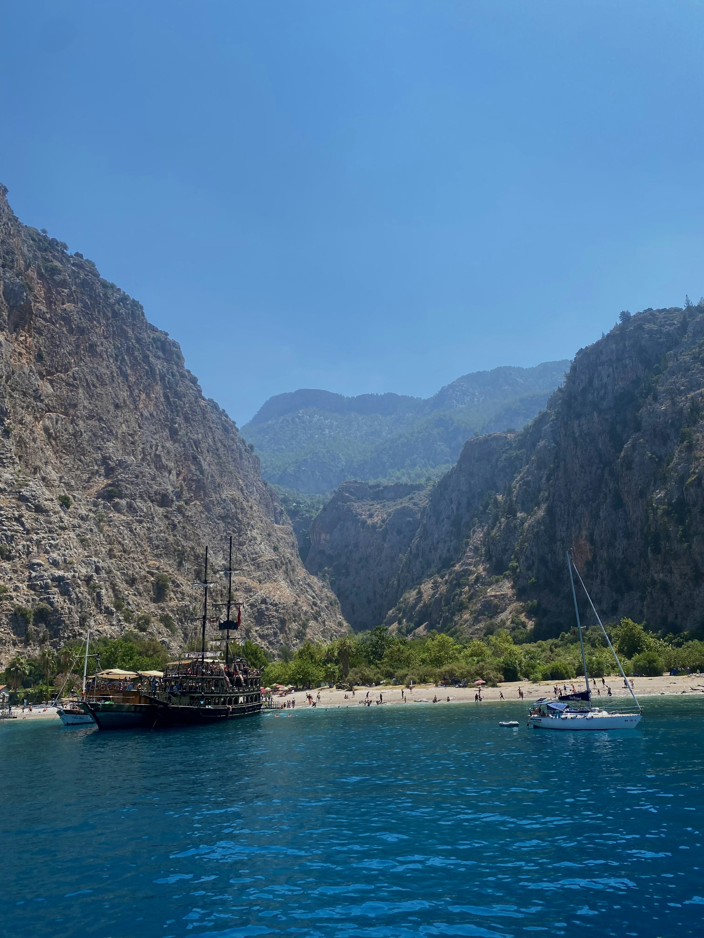 L'entrée de Butterfly Valley vue depuis la mer : de hautes falaises de chaque côté, une plage au fond, des bateaux ancrés dans l'eau turquoise.