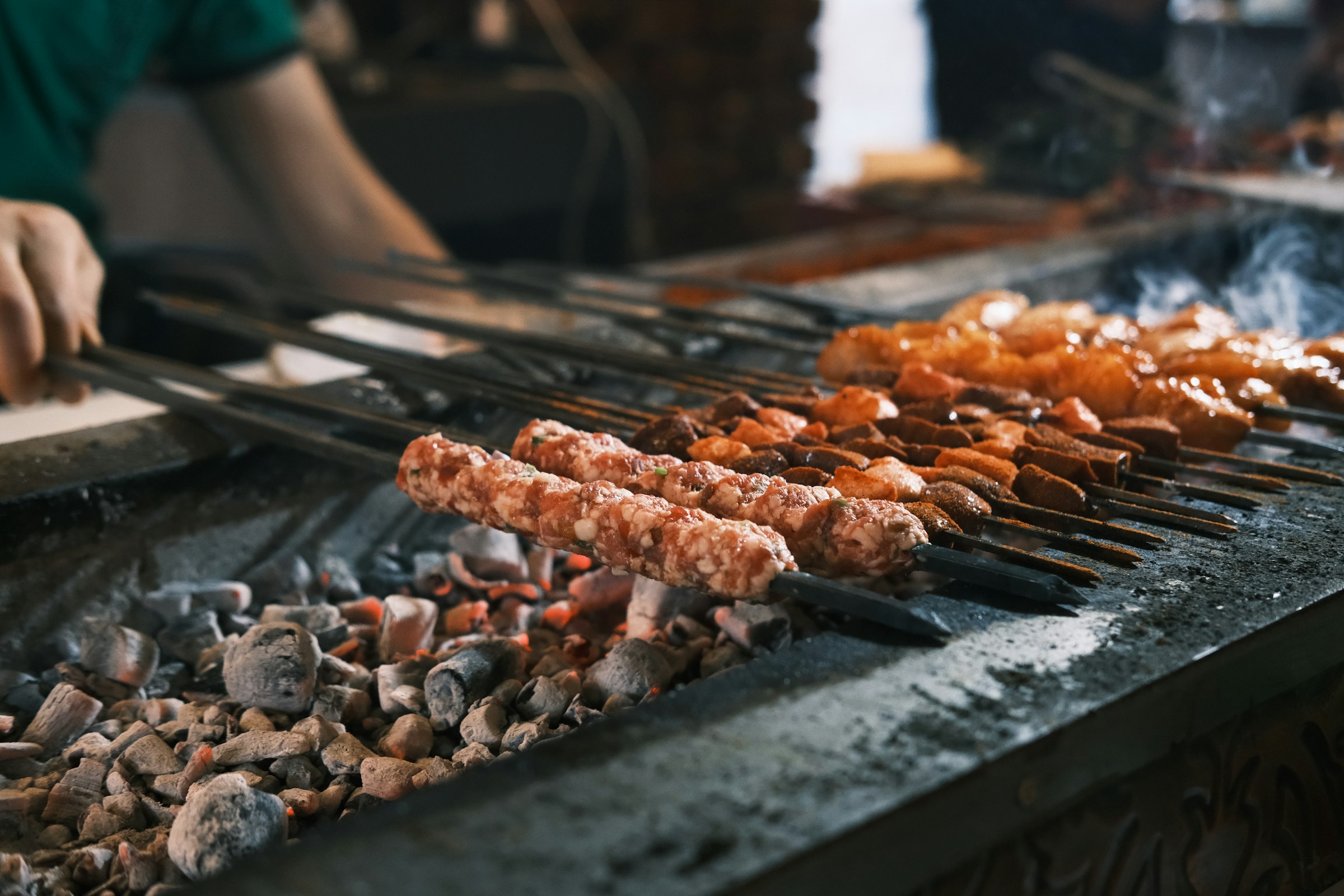Des brochettes turques qui grillent sur des braises rougeoyantes, une main qui les tourne à la pince, un filet de fumée.