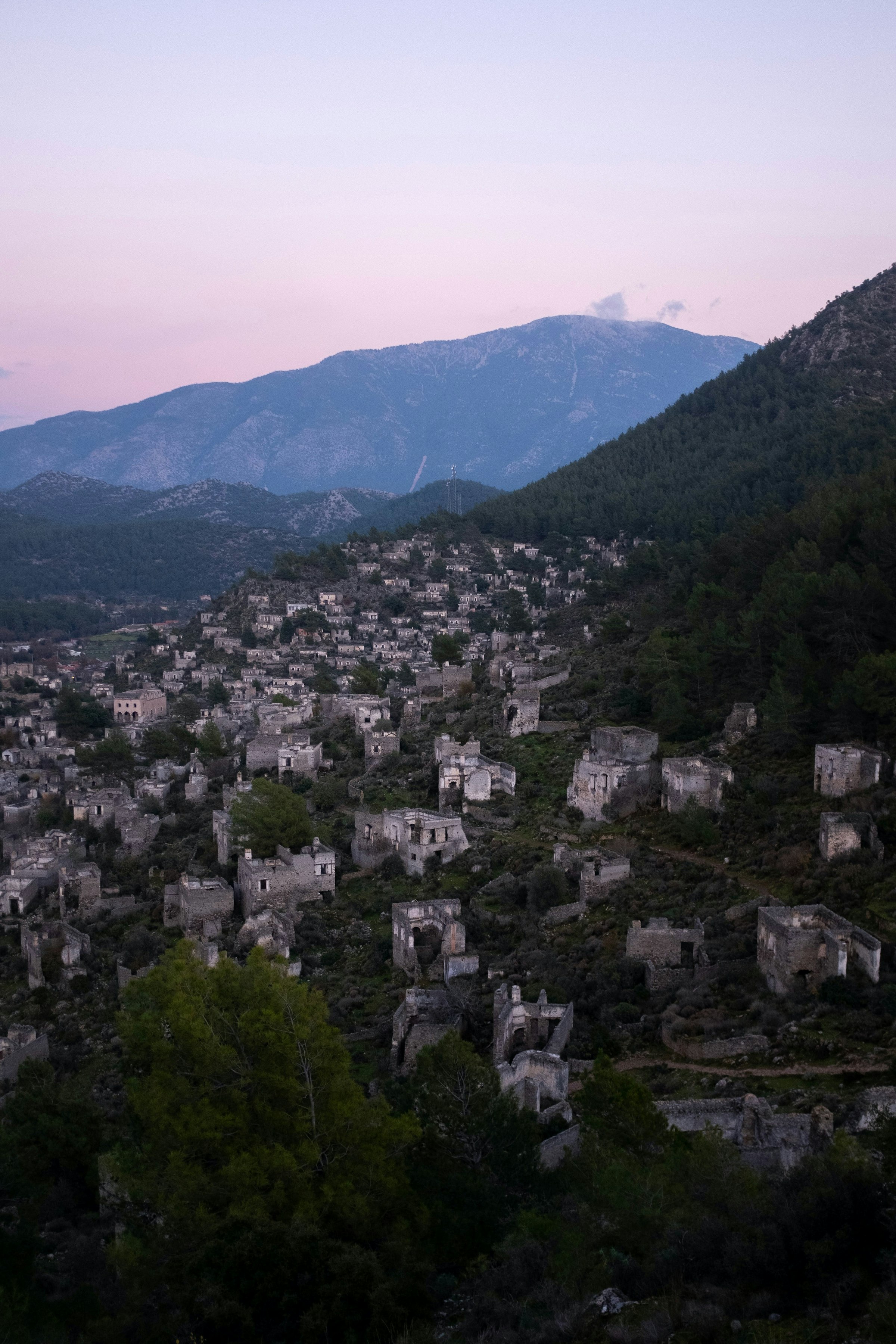 Le village fantôme de Kayaköy au crépuscule : des maisons de pierre en ruines étagées sur la colline boisée, un ciel rose, aucune personne.