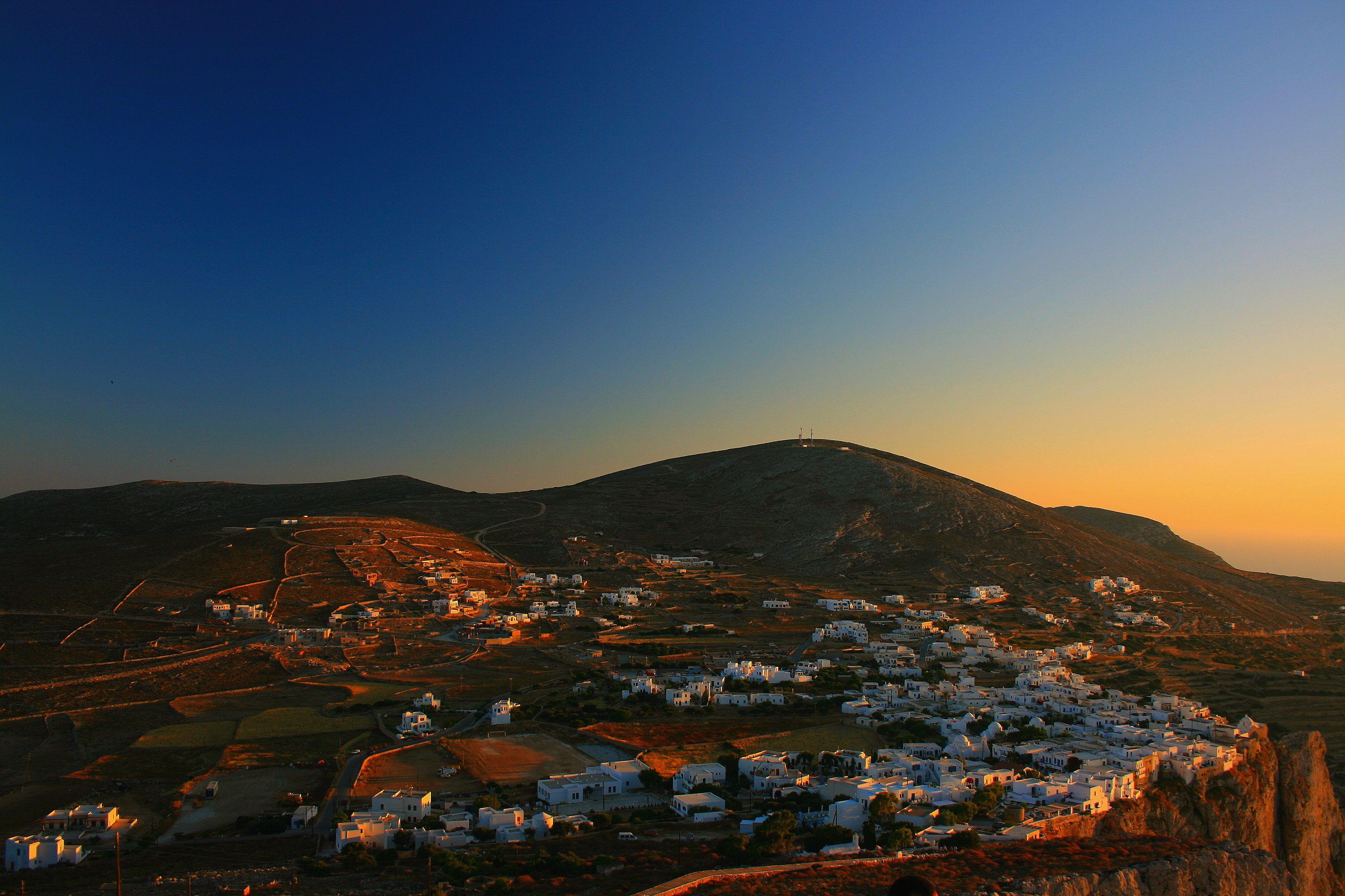 Le village de Chora de Folegandros au crépuscule : les maisons blanches groupées sur le flanc d'une colline aride, sous un ciel qui passe du bleu profond à l'orangé du couchant