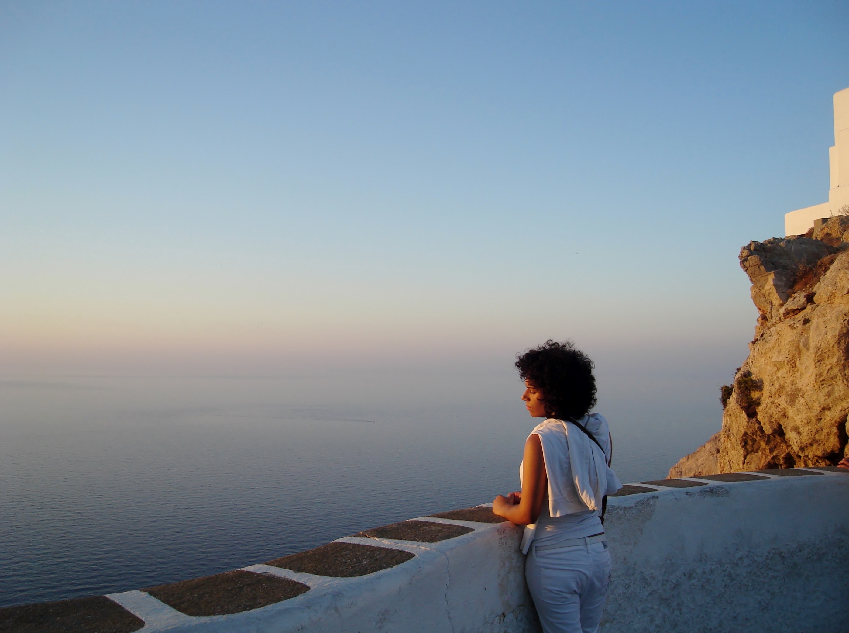 Une femme accoudée à un muret blanchi au bord de la falaise de Chora à Folegandros, regardant la mer immense au crépuscule, une chapelle blanche perchée sur le roc à droite