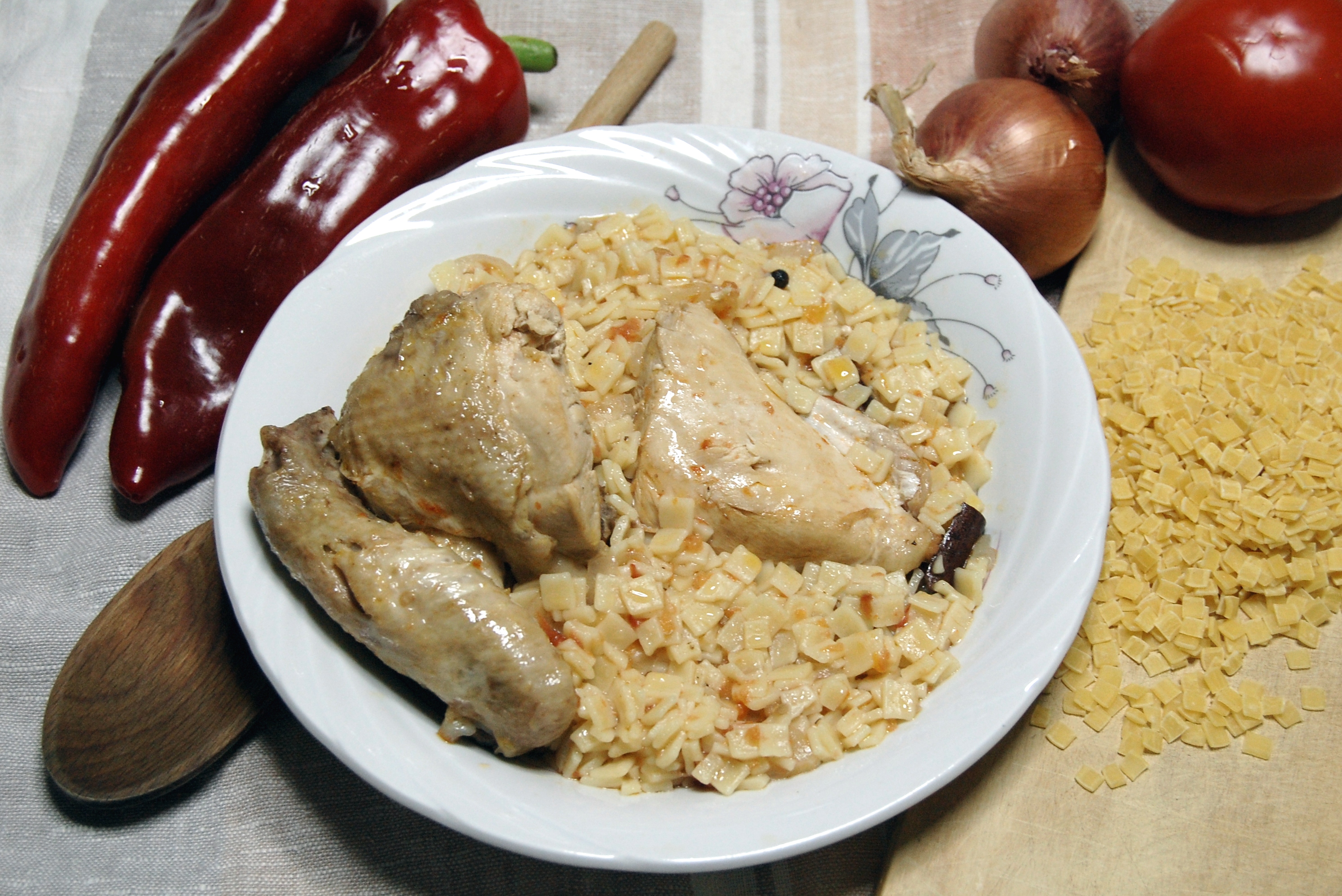 Une assiette de poulet aux hilopites (petites pâtes carrées coupées à la main) dans une sauce crémeuse, avec des poivrons rouges, des oignons et une tomate autour, et des pâtes crues à côté