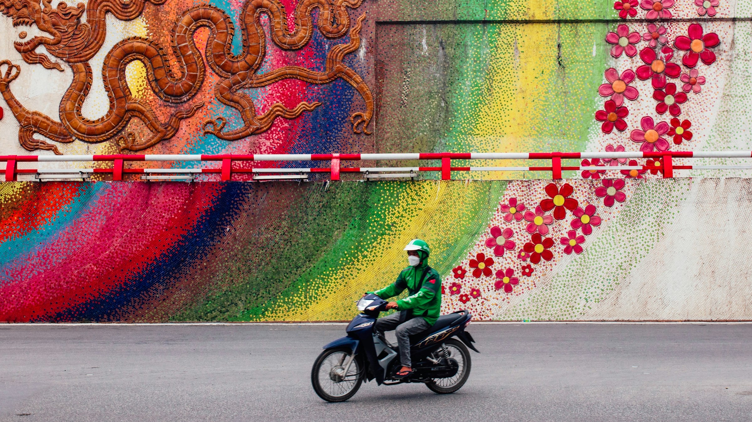 Livreur à moto (veste verte, masque) passant devant une immense fresque murale colorée — dragon et fleurs rouges sur fond arc-en-ciel, quartier Phúc Tân, Hanoi
