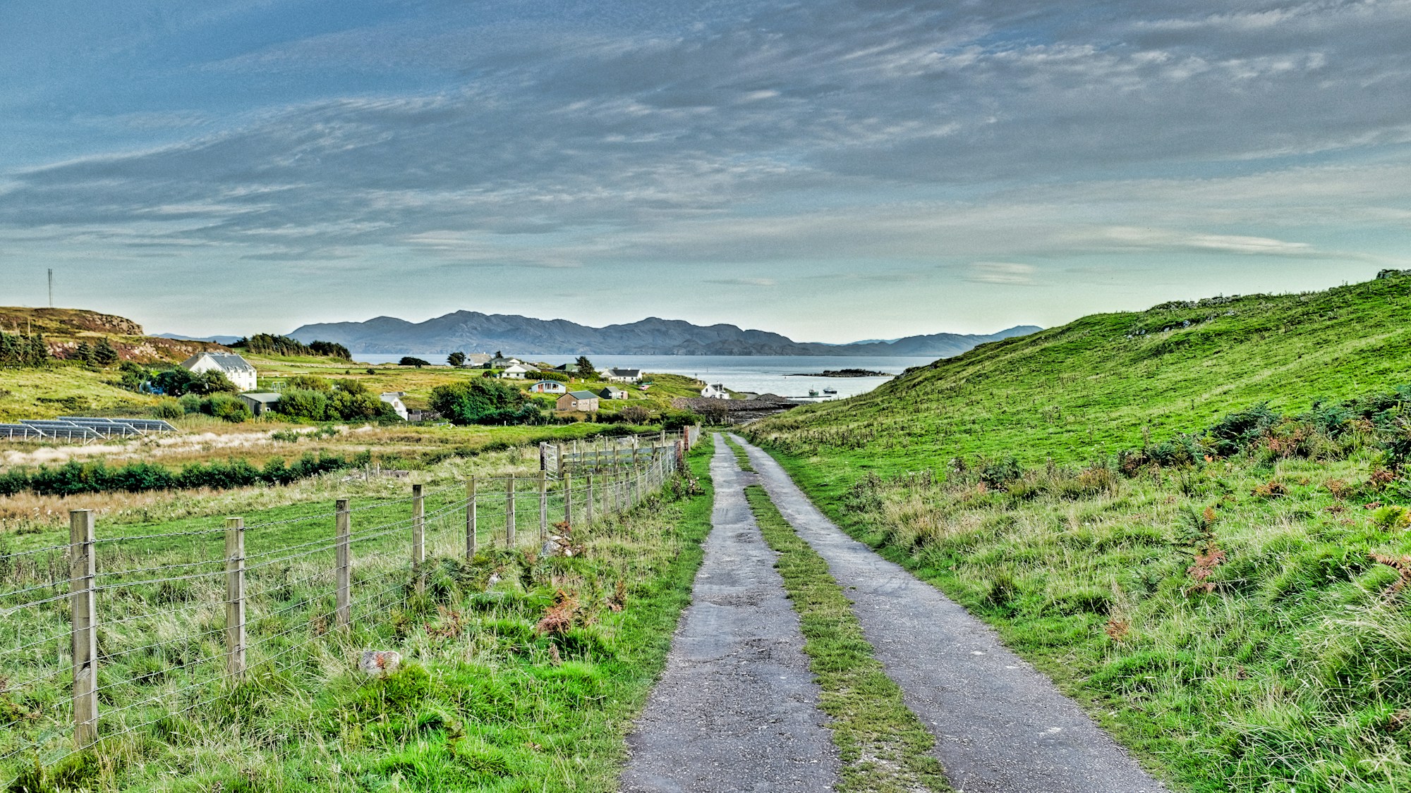 A narrow single-track lane, no centre line, running down between fenced fields to a scattering of white croft houses at the sea's edge, low hills across the water.