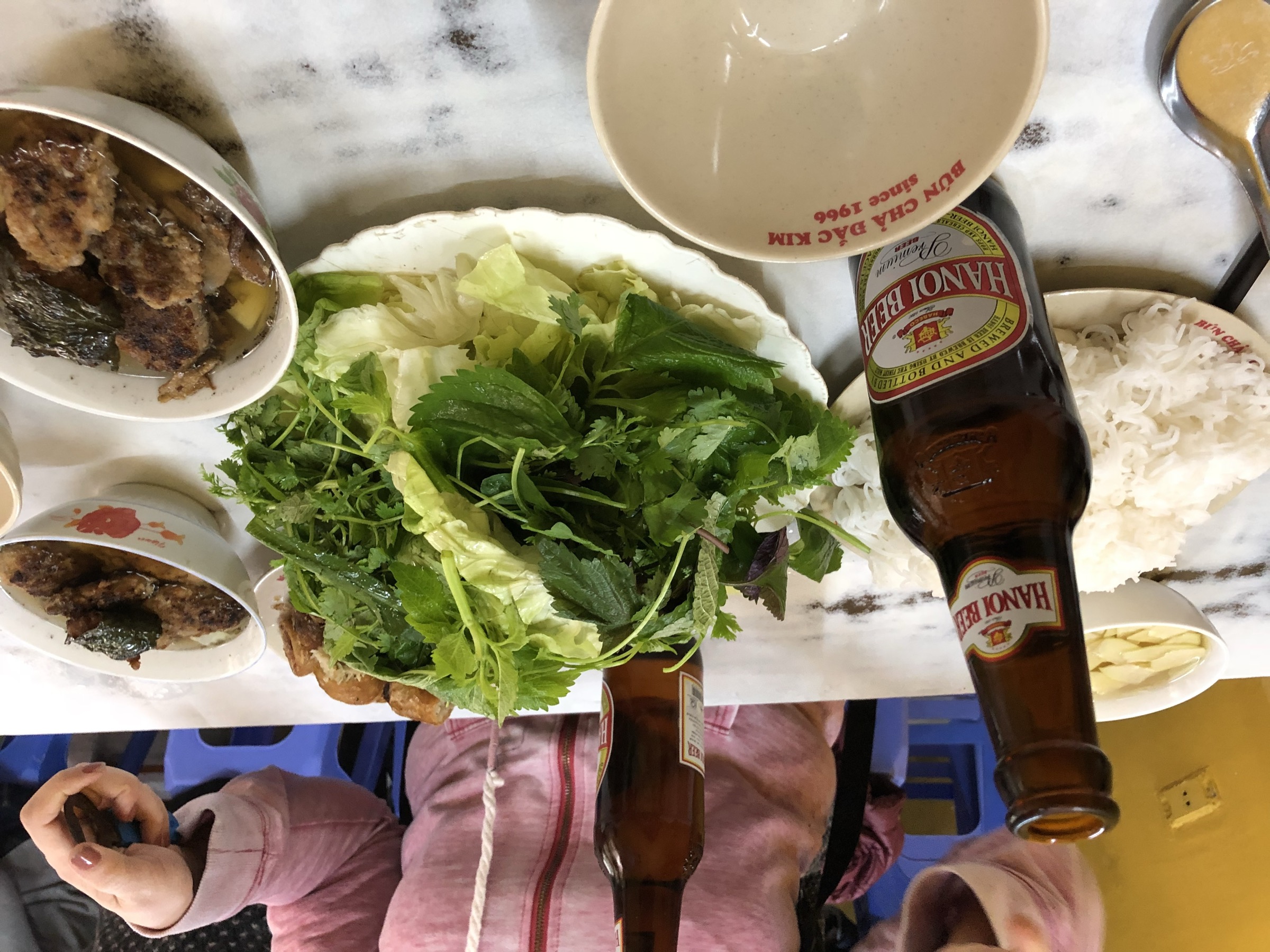 Plateau de bún chả à Hanoi — galettes de porc grillées dans un bol de bouillon, grande assiette d'herbes fraîches, vermicelles de riz et bière locale sur une table de marbre