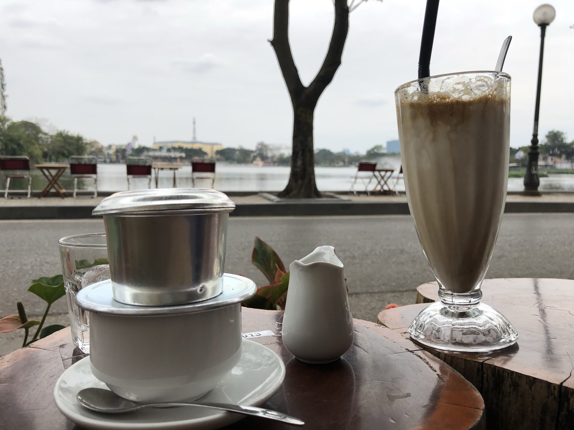 Verre de cà phê trứng (café à l'œuf) à la mousse crémeuse dorée et son filtre phin, sur une table donnant sur un lac de Hanoi