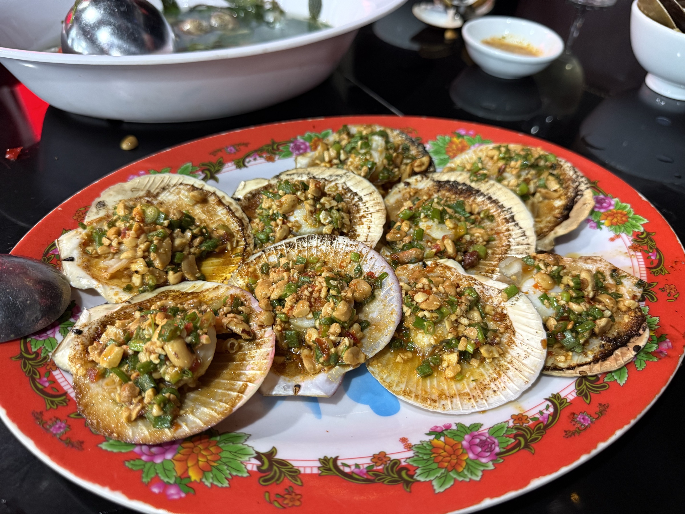 Coquilles Saint-Jacques grillées dans leur coquille, garnies d'ail haché, cacahuètes concassées et oignons verts, sur assiette à motifs floraux rouges avec coupelle de sauce — fruits de mer grillés de rue à Hội An