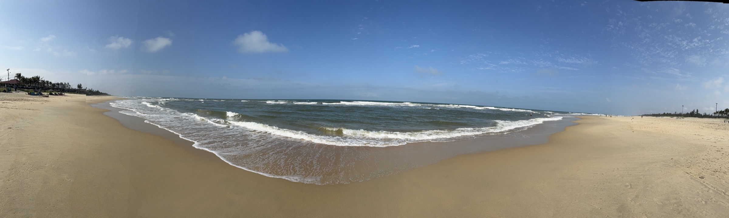 Plage de sable fin pratiquement déserte près de Hội An — vagues déferlantes blanches, rangée de palmiers et ciel bleu avec quelques nuages