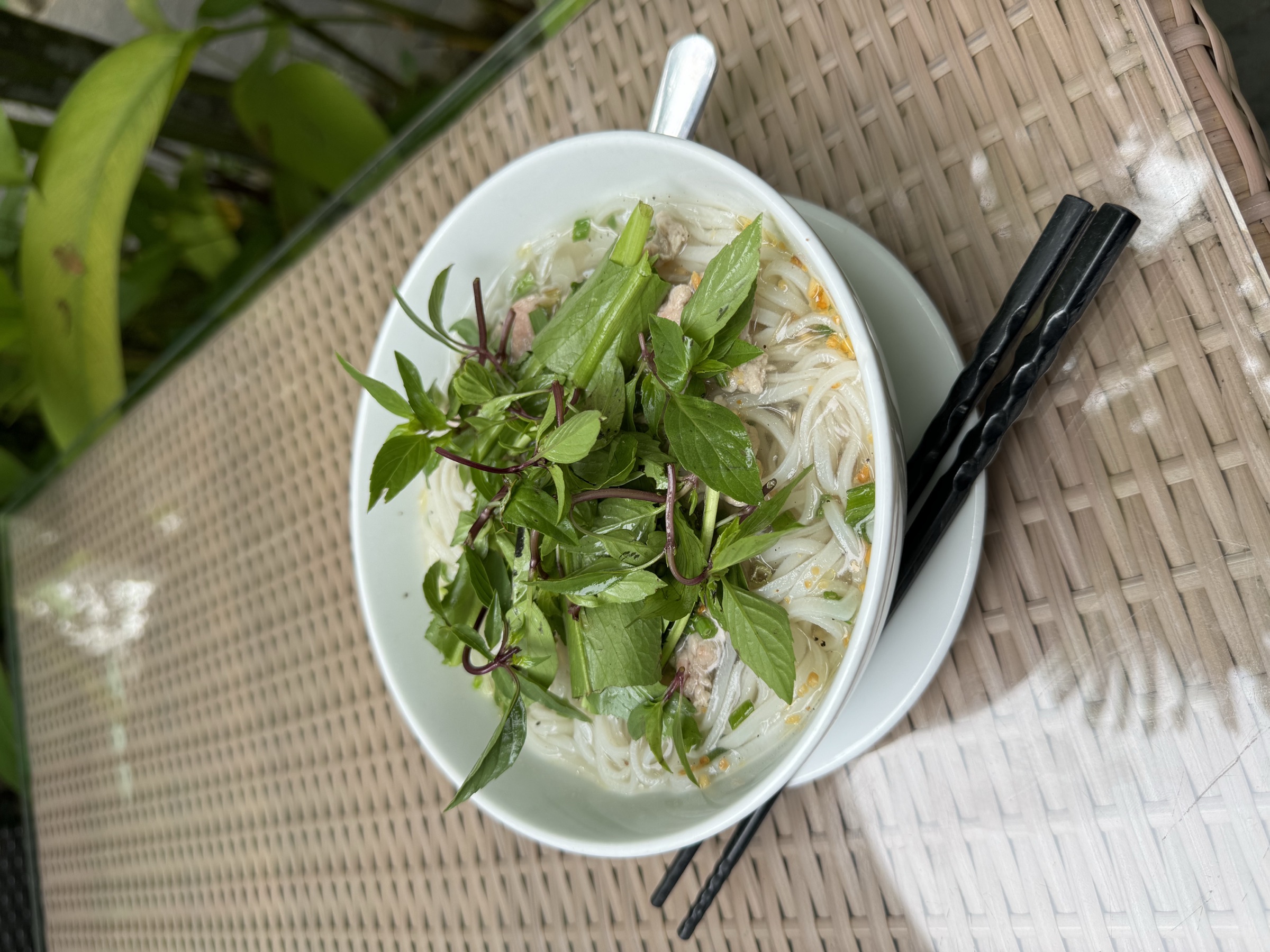Bol de soupe de nouilles de riz abondamment garni de feuilles d'herbes fraîches entières — basilic thaï et menthe, baguettes noires posées sur table en rotin en plein air