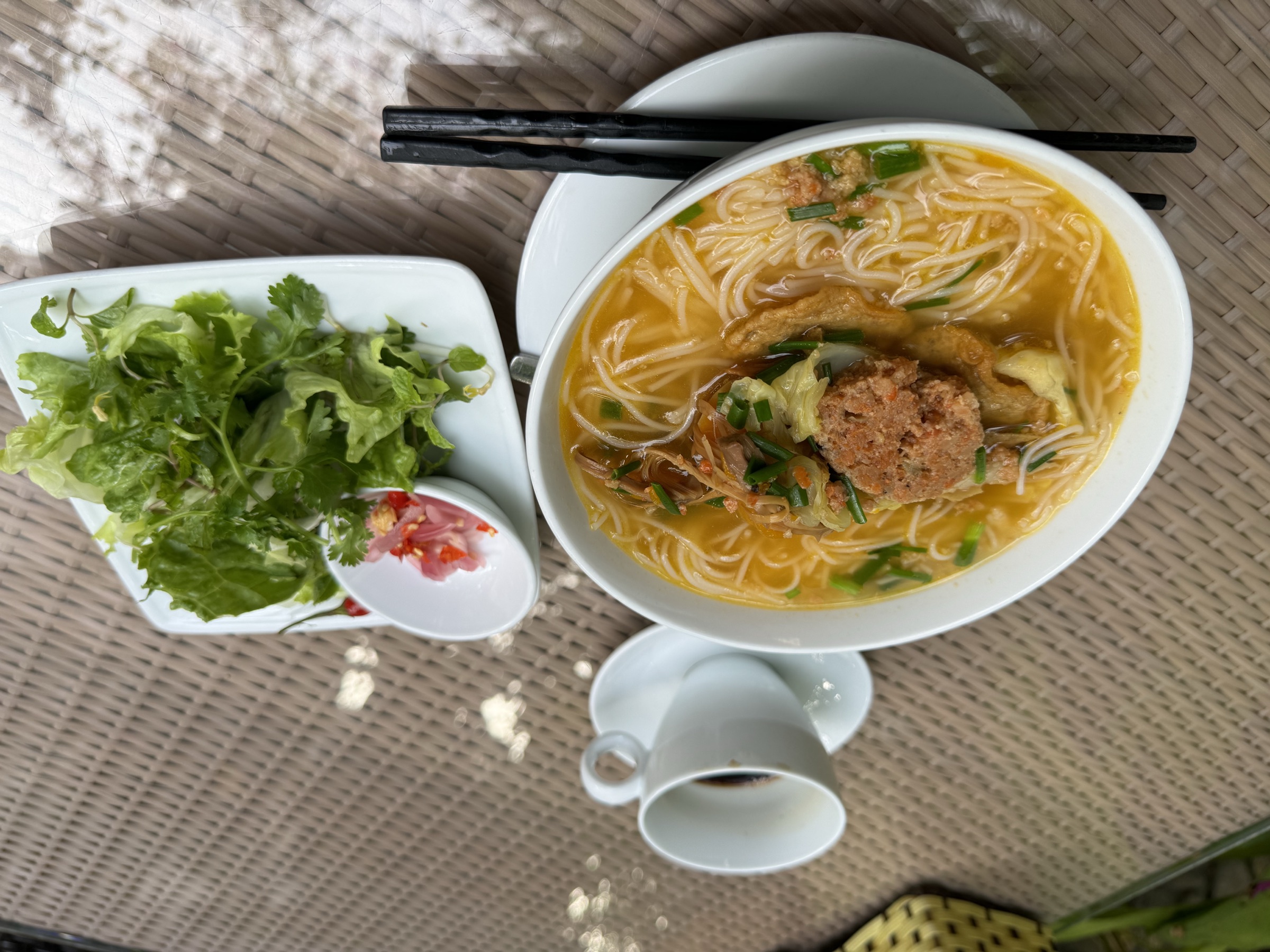 Bol ovale de soupe de nouilles au bouillon doré garni de tofu frit, boulette de viande et oignons verts, accompagné d'une petite salade fraîche et piments rouges, sur table en rotin