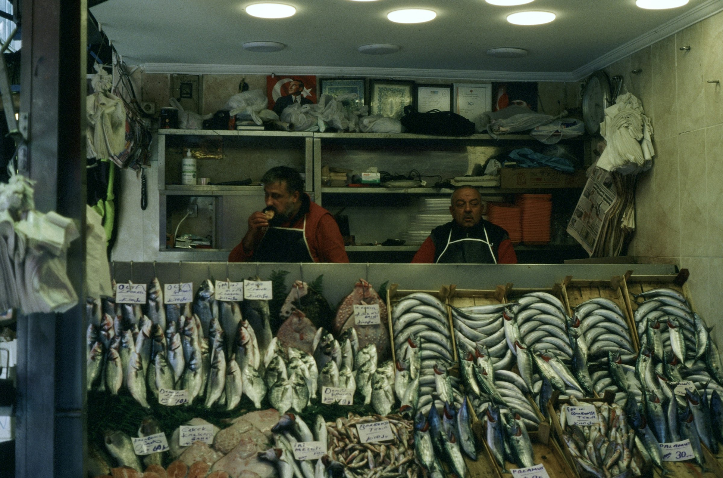 Deux poissonniers derrière leur étal couvert de poissons frais dans un marché d'Istanbul, un drapeau turc au fond.