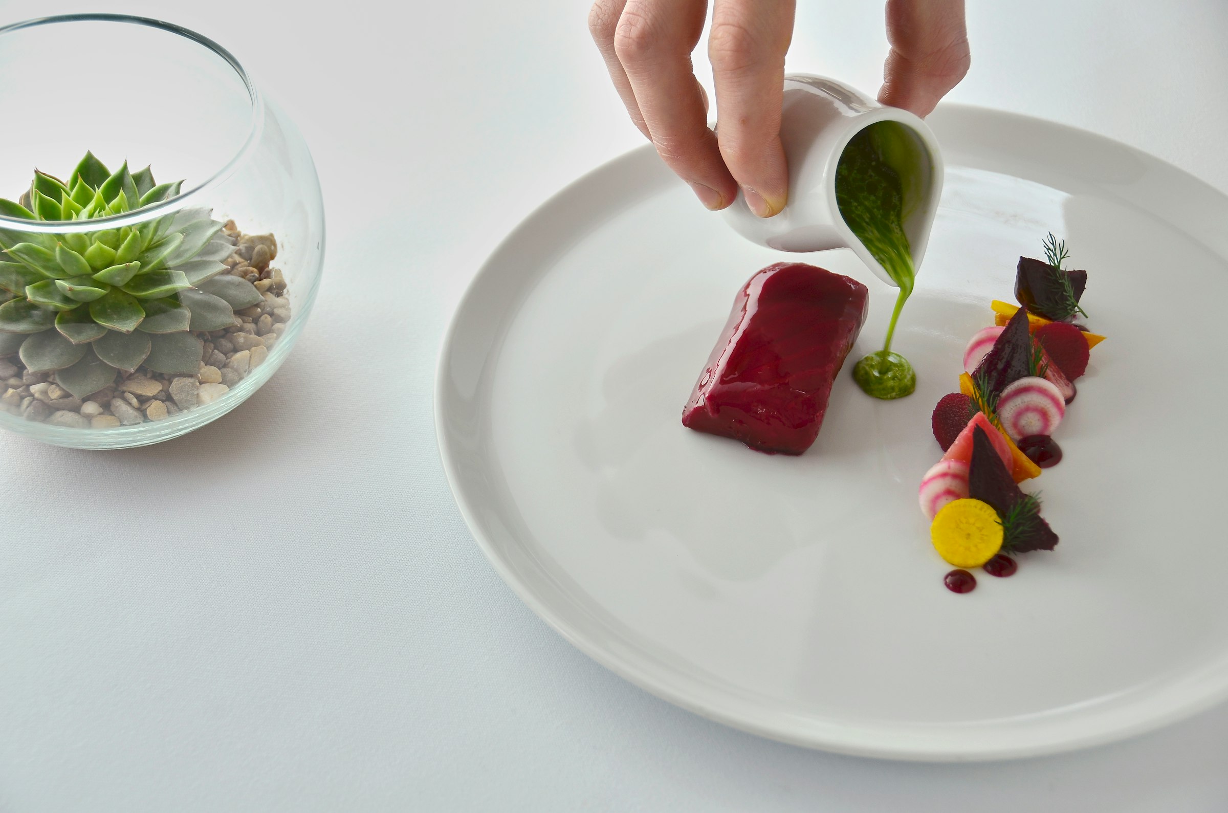 Une main verse une sauce verte sur une assiette gastronomique épurée — betterave glacée, légumes-racines, dressage soigné sur fond blanc.