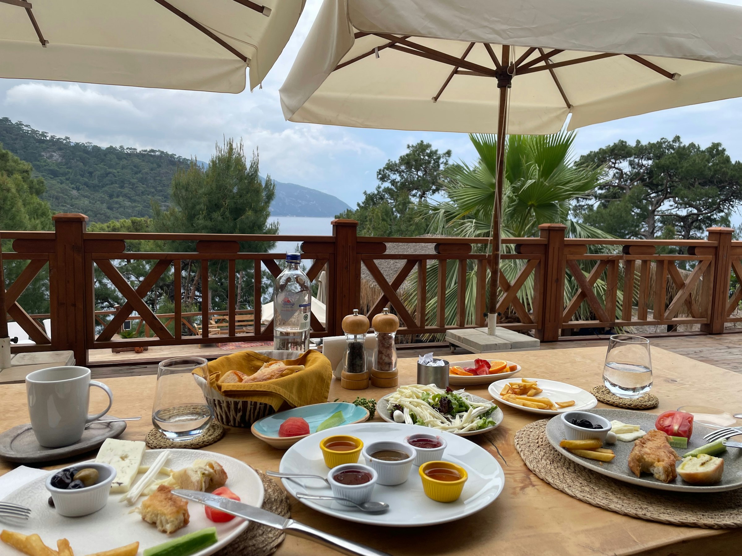 Une table de petit-déjeuner turc — fromages, olives, œufs, confitures, pain — sur une terrasse en bois, la mer et les collines en arrière-plan.