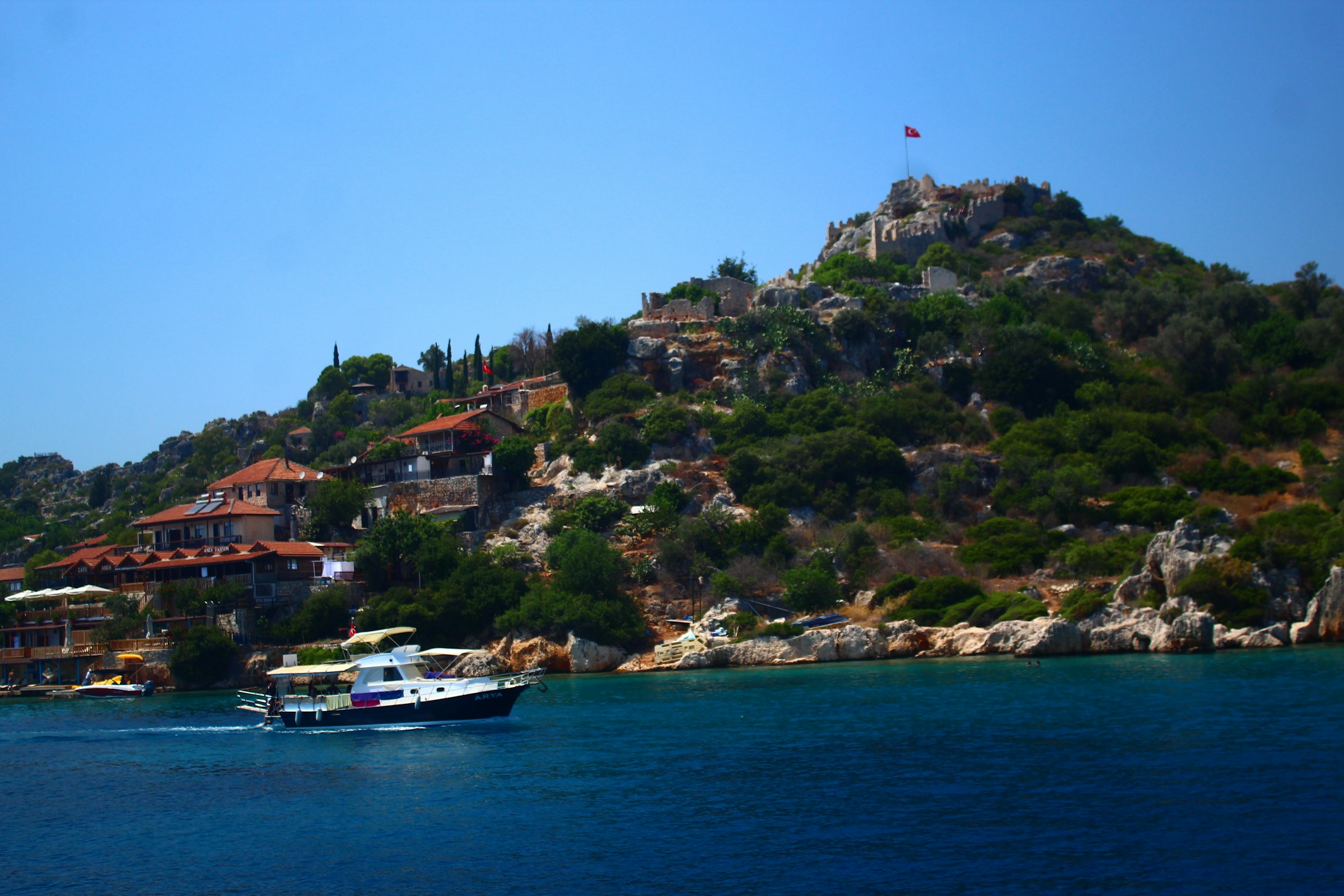 Un bateau longe la colline de Simena (Kekova) : maisons à toits rouges, le château au sommet avec un drapeau turc, l'eau turquoise au premier plan.
