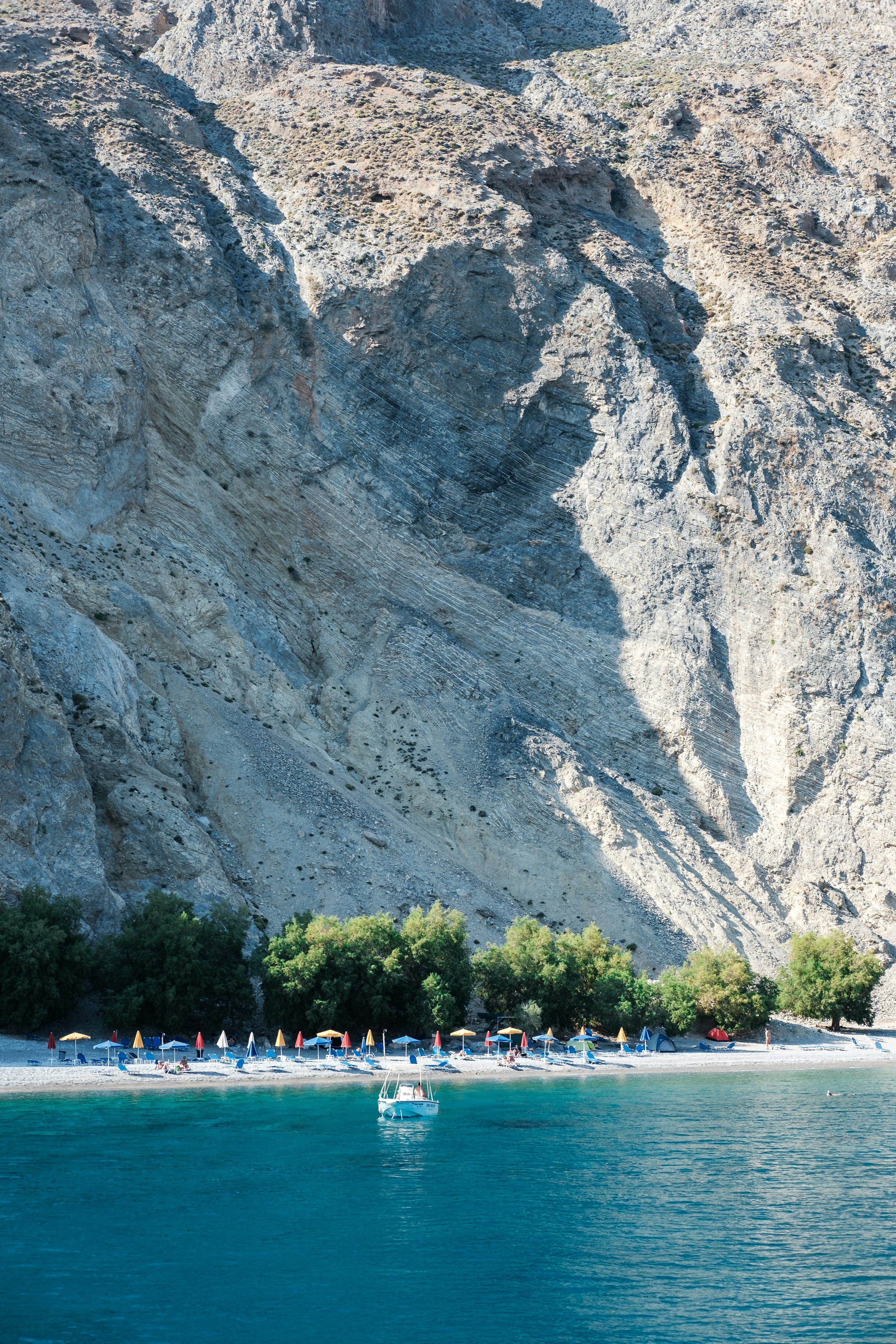 Glyka Nera (Sweetwater Beach), près de Loutro : une plage de galets au pied d'une immense falaise calcaire pâle, des tamaris nourris par les sources d'eau douce, une barque et des parasols dans une eau turquoise.