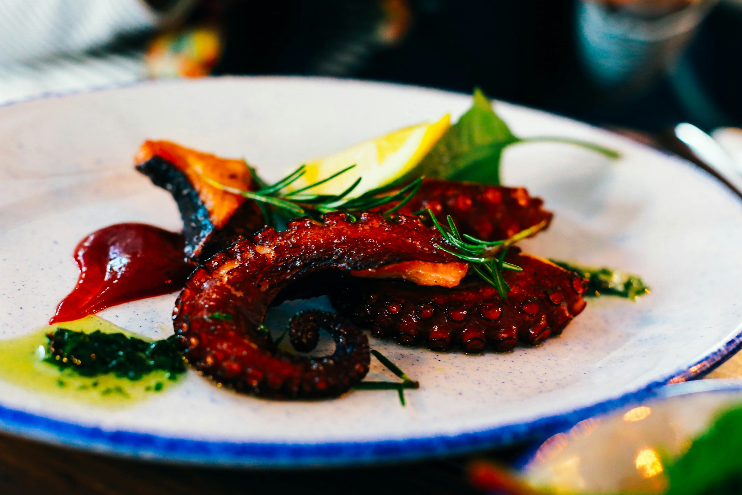 Une assiette de poulpe grillé : des tentacules charbonnés et glacés, un quartier de citron, des brins de romarin et des touches d'huile verte et de sauce, sur une assiette en céramique au liseré bleu, dans une ambiance de taverne.