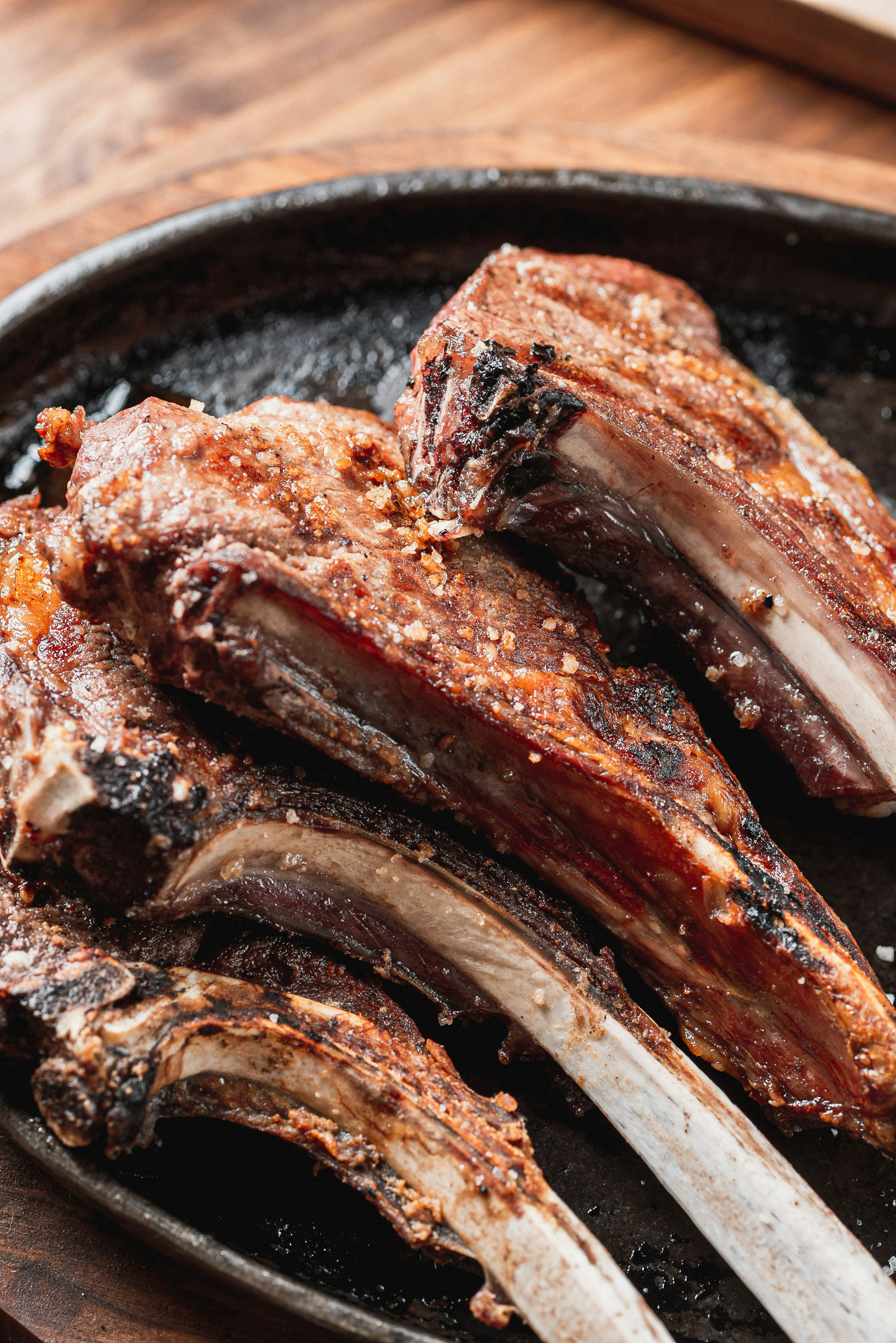 Des côtelettes d'agneau grillées (paidakia), os à l'air et croûte d'épices dorée et caramélisée, dans une poêle en fonte noire sur une table de bois.
