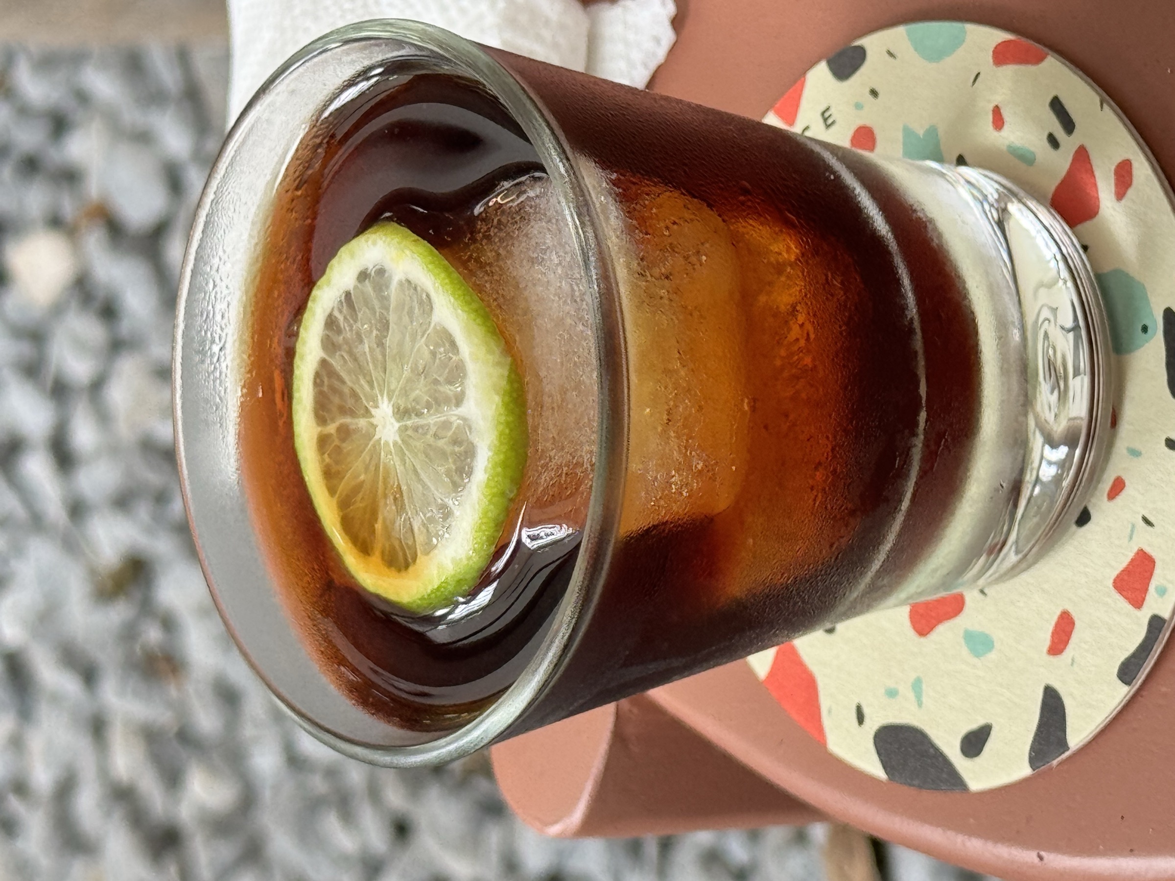 Verre de cà phê đá — café glacé vietnamien brun foncé avec rondelle de citron vert, posé sur un sous-verre en terrazzo coloré, lumière naturelle douce