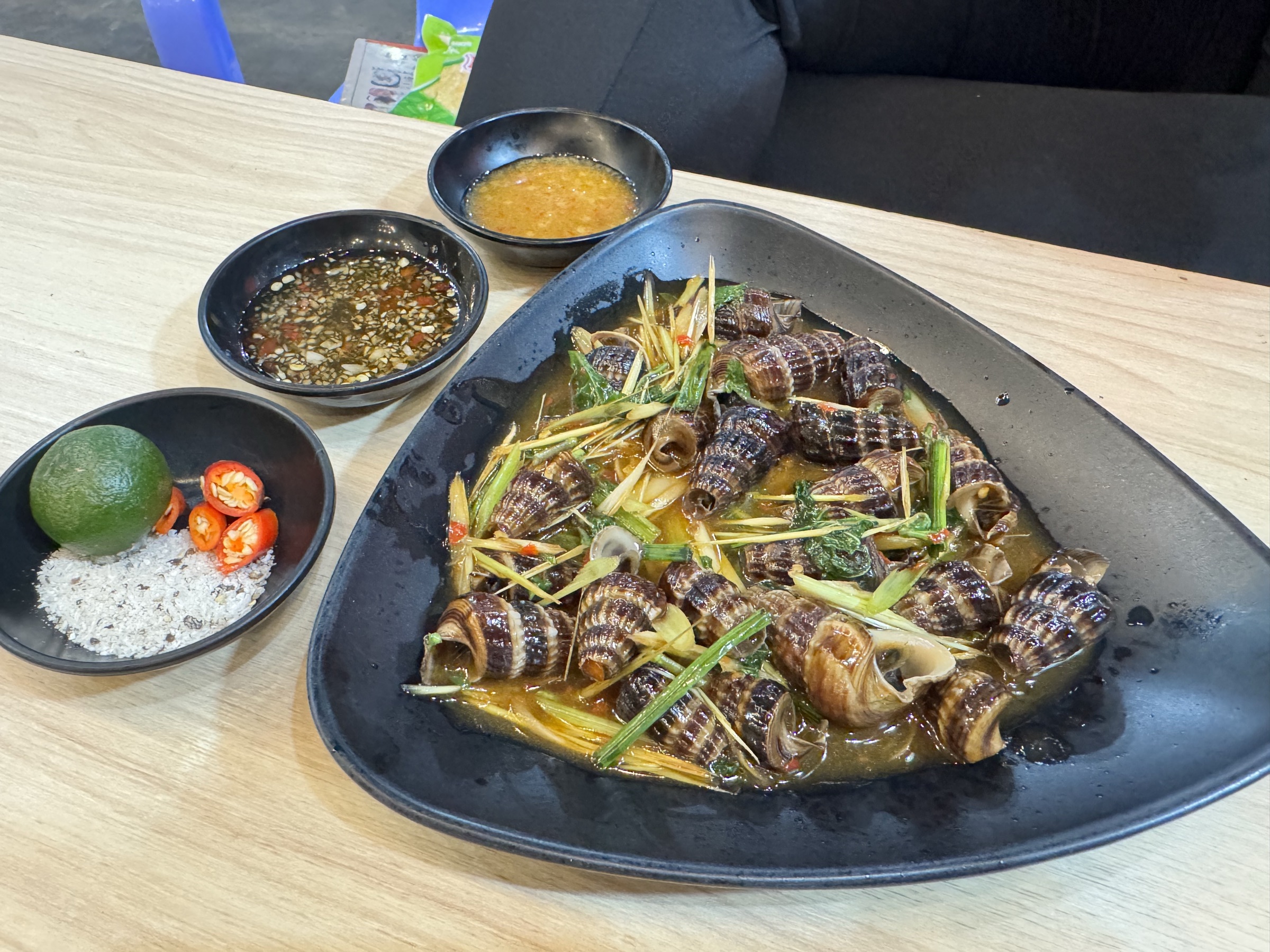 Plateau d'ốc (coquillages de mer) cuits dans une sauce brune à la citronnelle et aux piments, présenté dans un plat noir triangulaire avec trois coupelles d'accompagnement (nuoc-mâm, sauce épicée, citron vert et sel), rue des escargots de Quy Nhơn