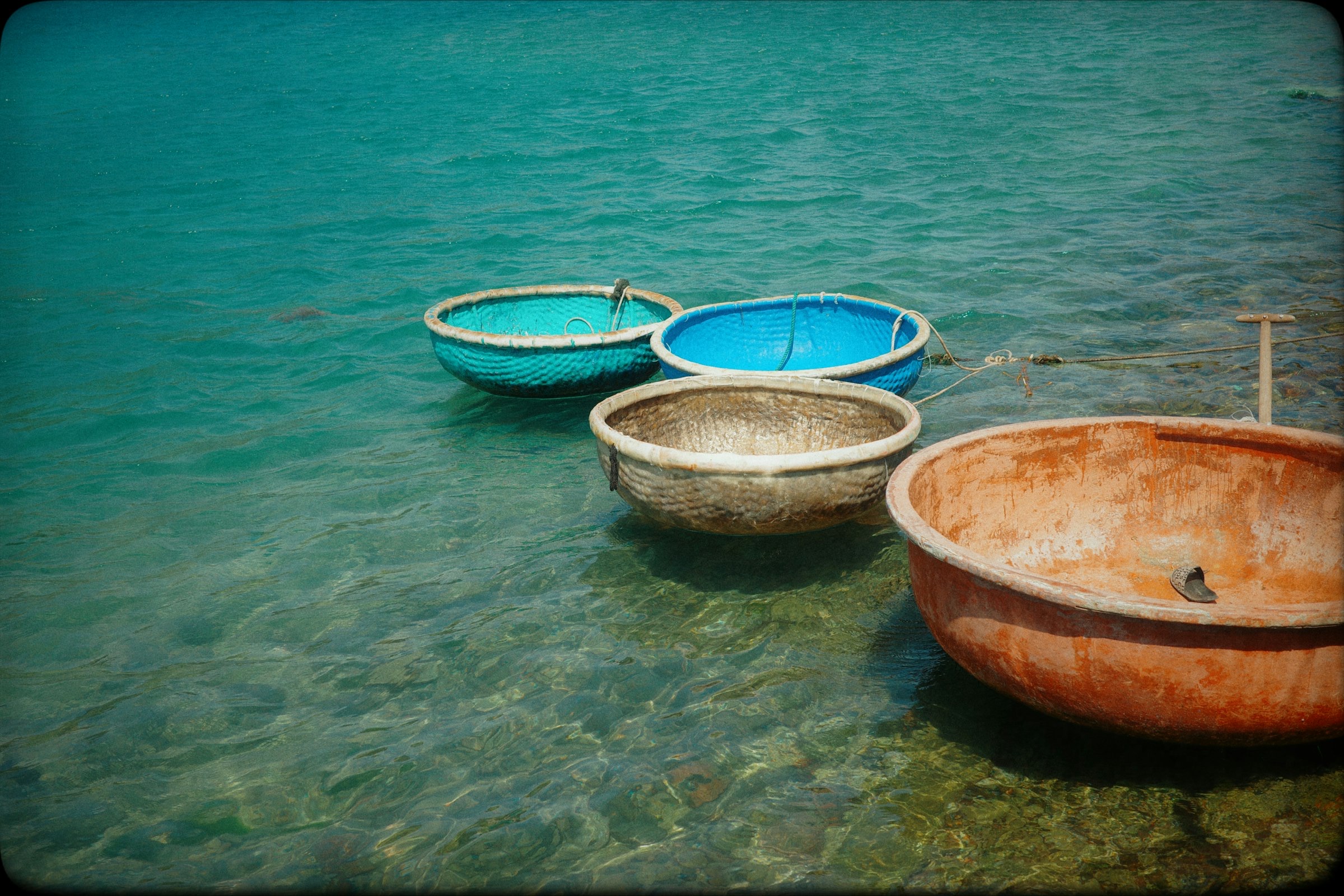 Trois coracles ronds (thuyền thúng) — turquoise, bleu et brun rouille — amarrés dans une eau bleu-vert cristalline à Bãi Xếp, composition forte avec légère patine vintage