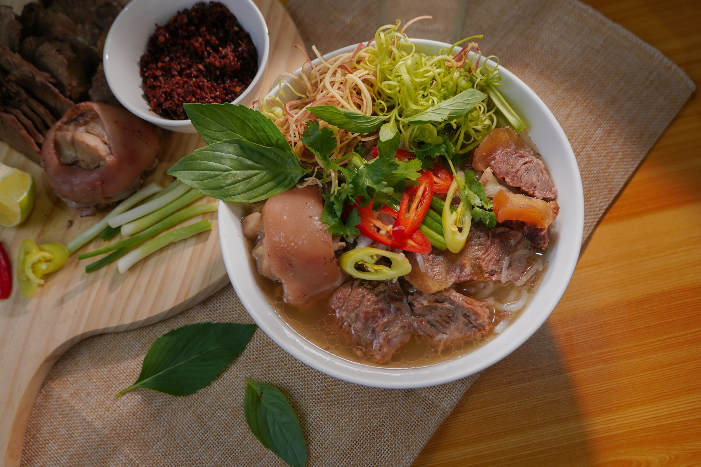 Bol de soupe de nouilles vietnamienne — vermicelles, tranches de viande de porc et bœuf, herbes fraîches (basilic, coriandre, pousses de soja), piments rouges et verts dans un bouillon brun, mis en scène sur planche de bois avec épices et citron vert