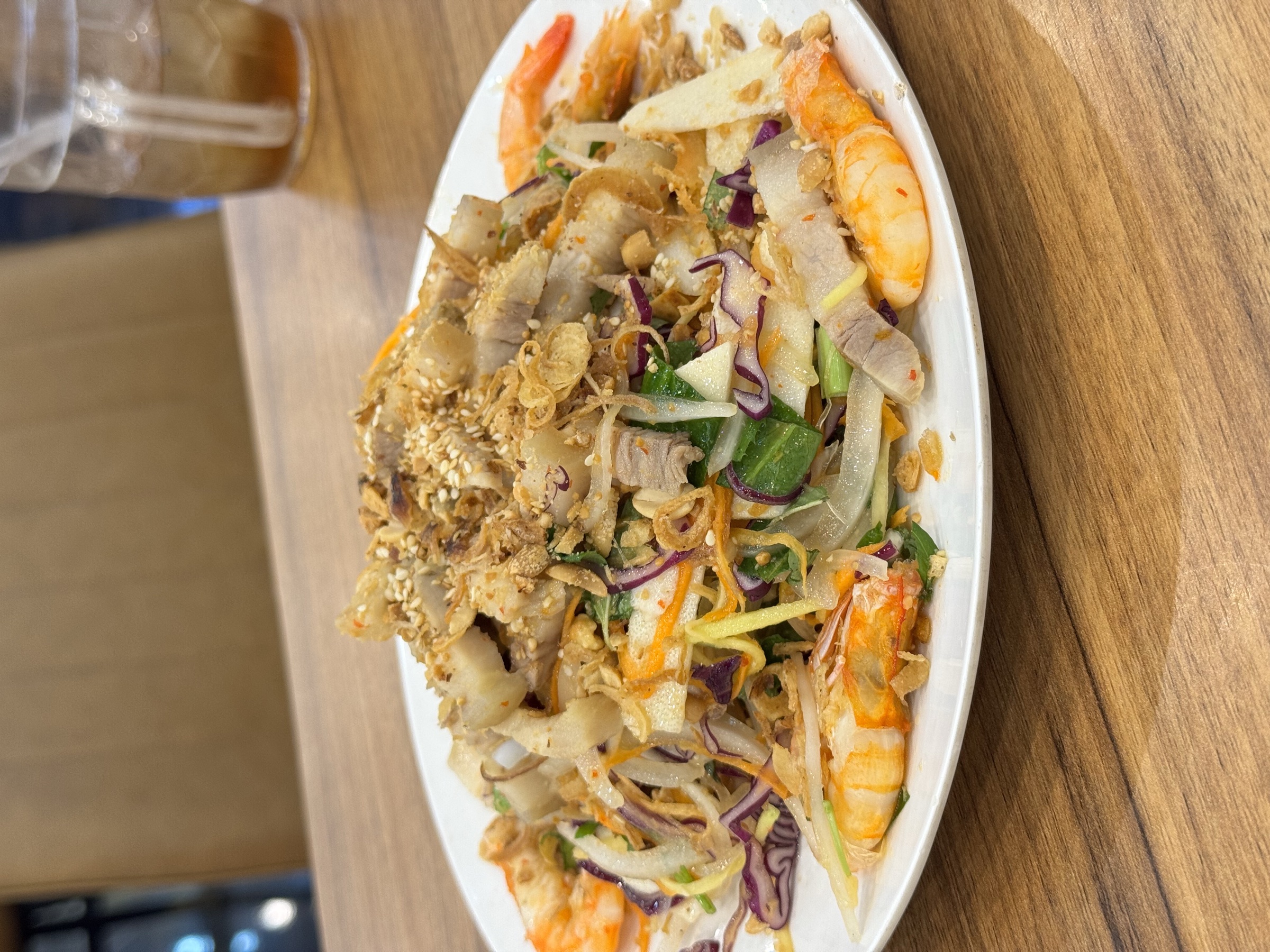 Assiette ovale de gỏi vietnamien aux crevettes — chou, carottes, herbes et condiments, photographiée en légère plongée sur table en bois, restaurant local de Quy Nhơn