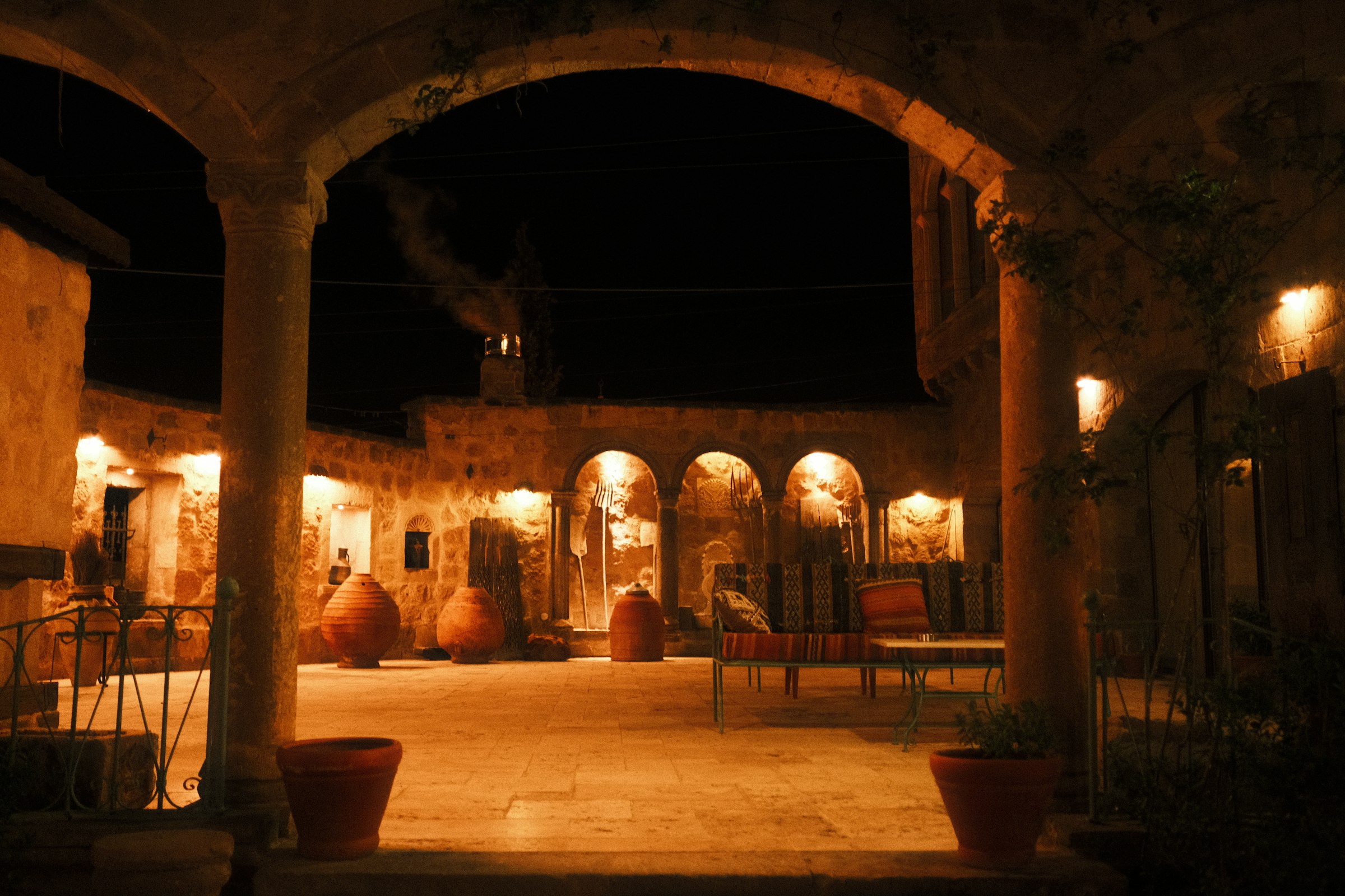 Un ancien patio en pierre la nuit : des colonnes et des arches voûtées éclairées par des lanternes ambrées, de grandes jarres en terre cuite, des tables en fer forgé.
