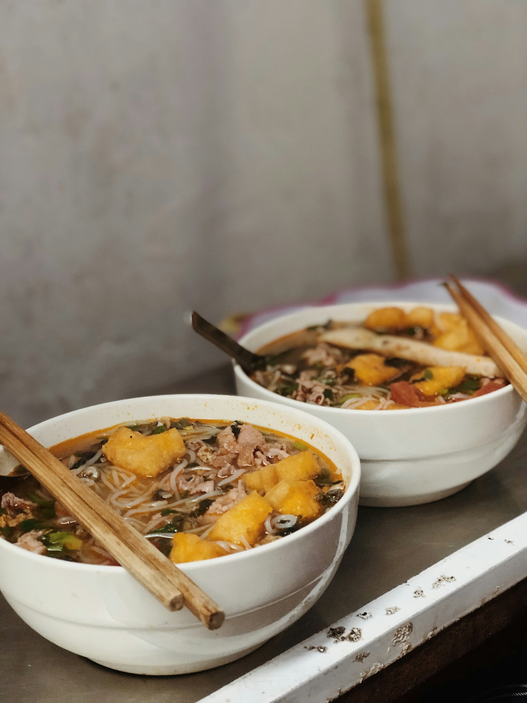 Deux bols de bún au bouillon orangé-rougeâtre — tofu frit doré, baguettes posées sur les rebords, servis sur plateau en inox dans une cantine de rue à Saïgon