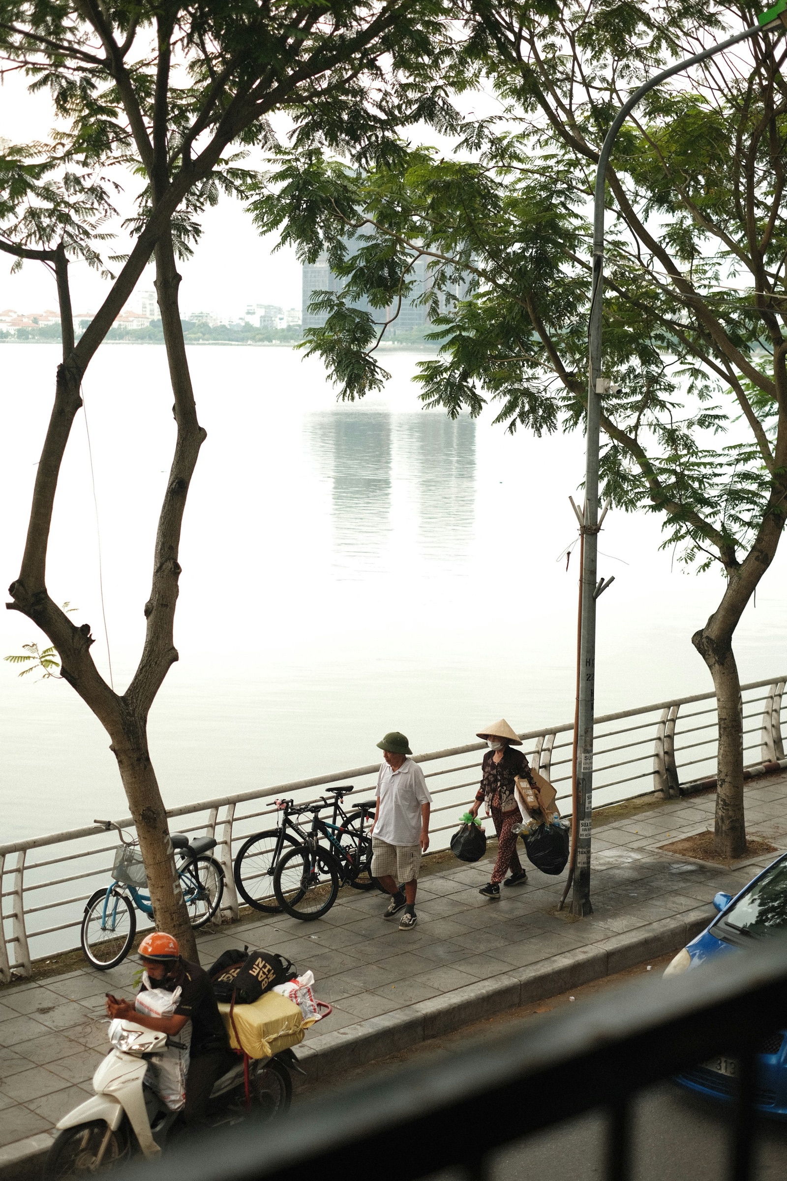 Promenade arborée au bord de l'eau à l'aube — passants, chapeau conique et vélos le long de la berge