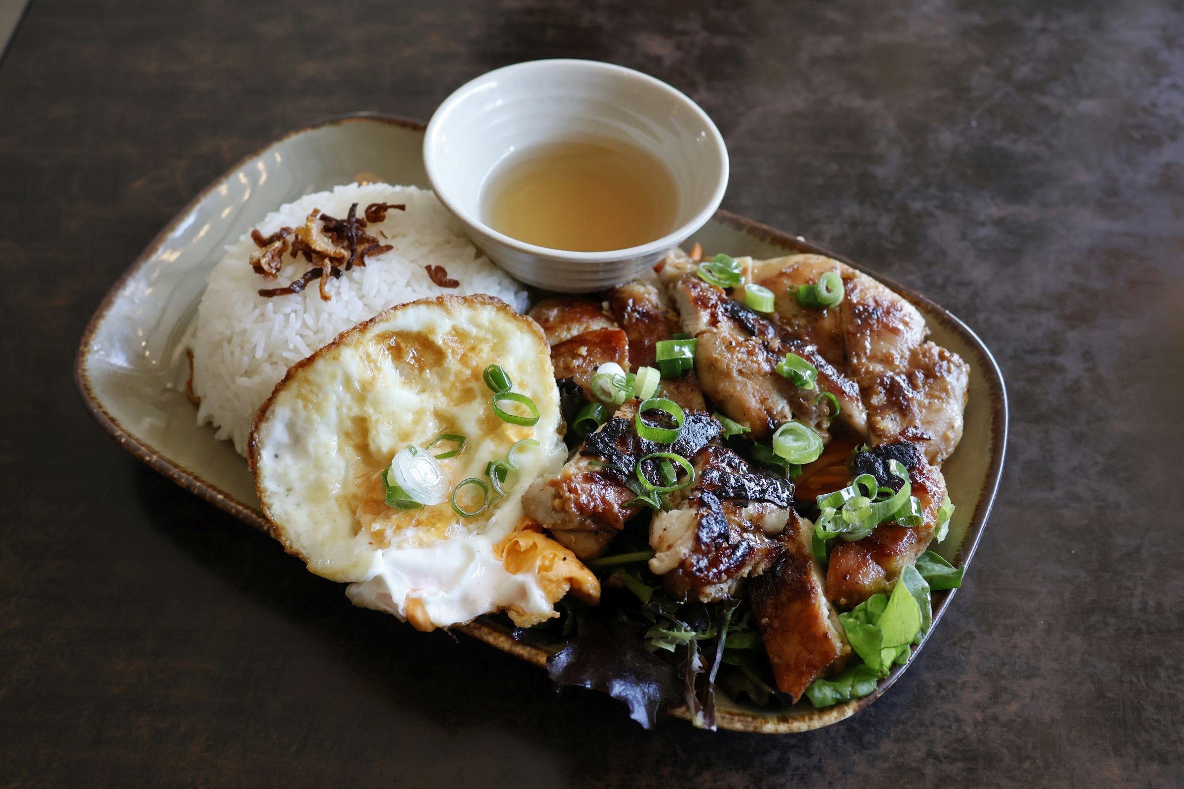 Assiette de cơm tấm — riz brisé avec viande grillée dorée garnie d'oignons verts, œuf au plat et bol de sauce à côté, lumière naturelle