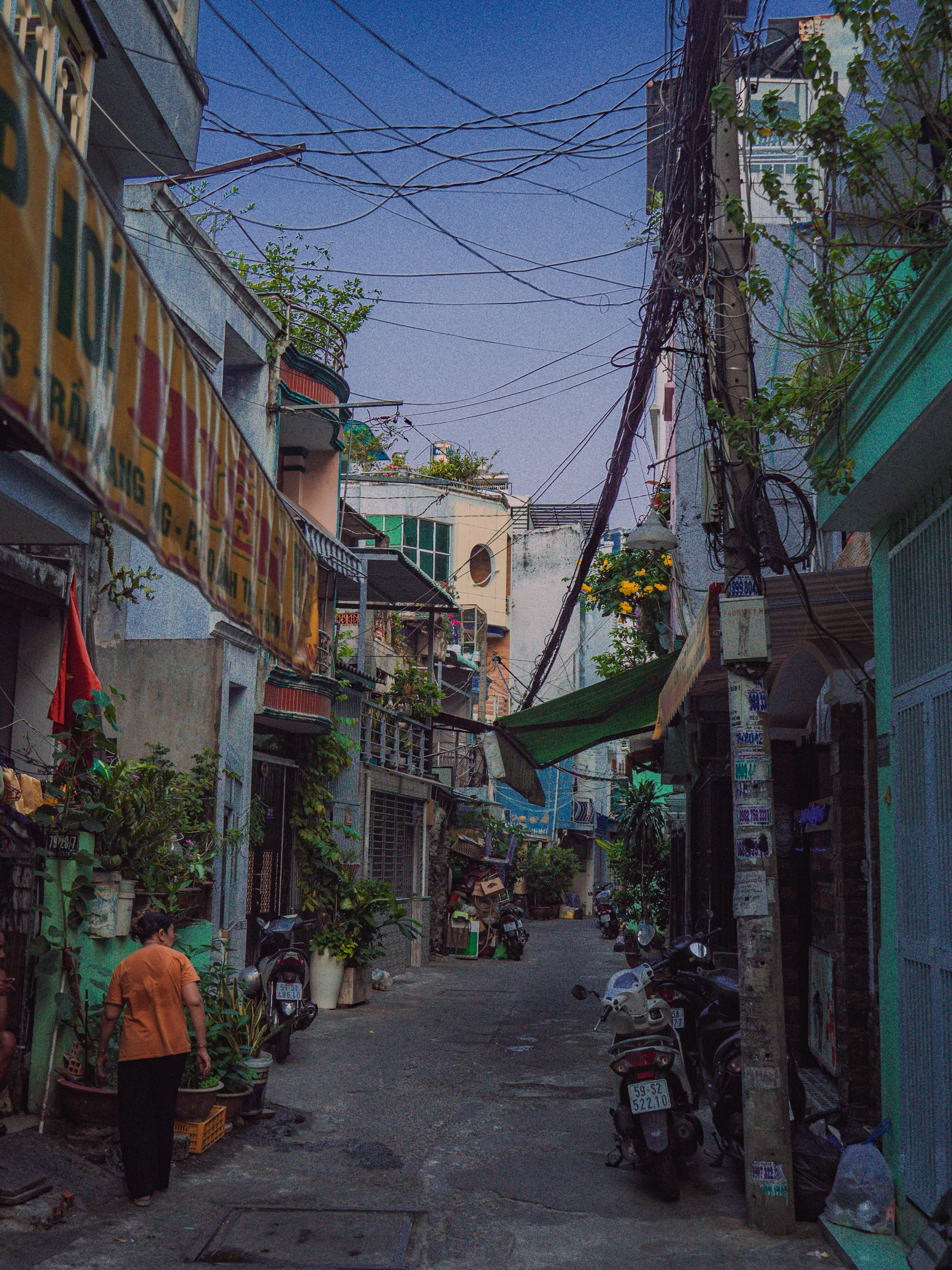 Ruelle (hẻm) étroite à Saïgon — immeubles serrés, fils électriques enchevêtrés, motos garées et un passant en t-shirt orange, ciel bleu dégagé