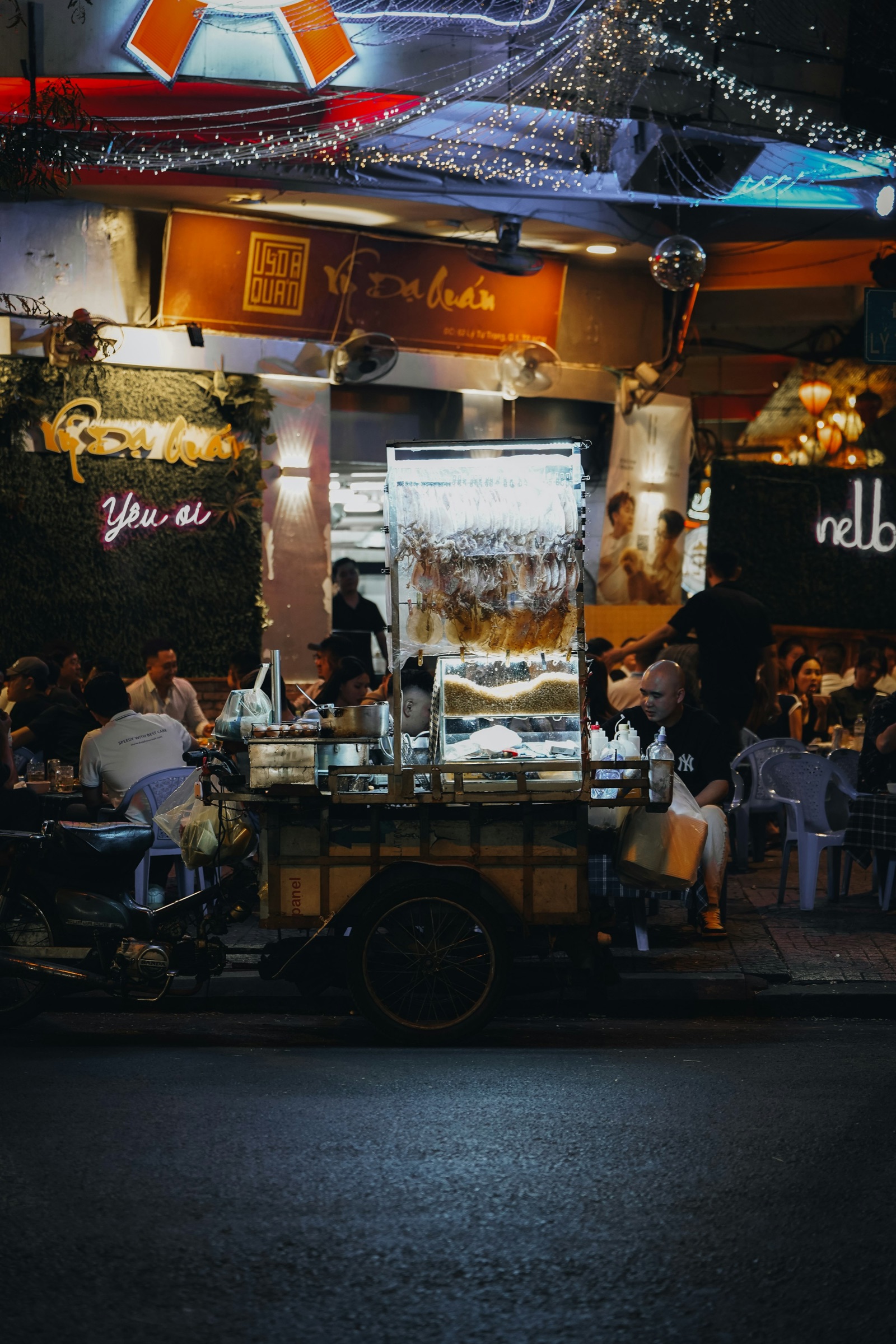Vendeur ambulant de fruits de mer la nuit à Saïgon — chariot vitré contenant des escargots et coquillages, néons de restaurants en arrière-plan, foule attablée dans la rue