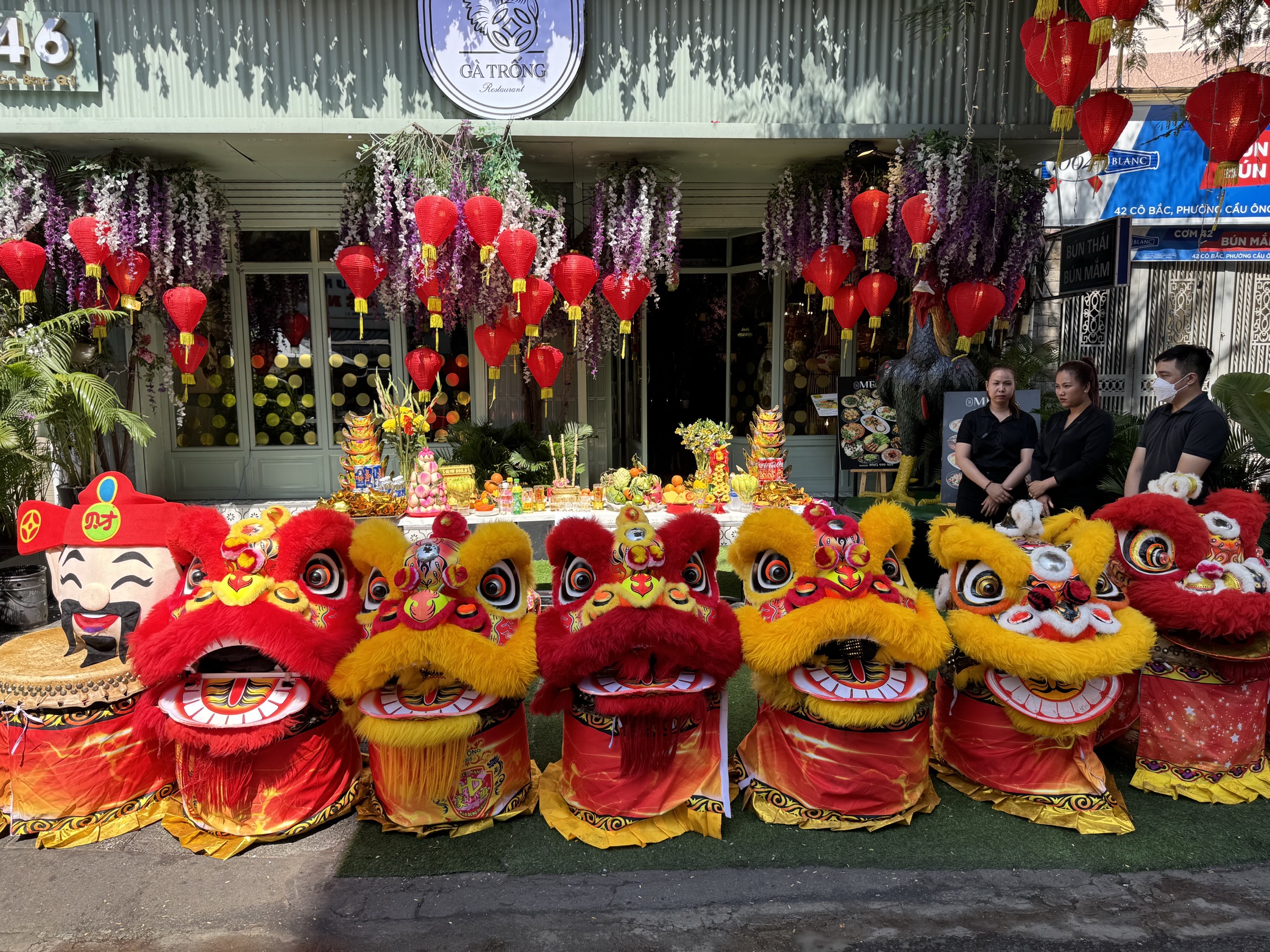 Têtes de lions de danse rouge et jaune alignées devant un restaurant aux lanternes rouges et glycines violettes — animation de rue pour le Tết à Saïgon, membres du personnel en noir