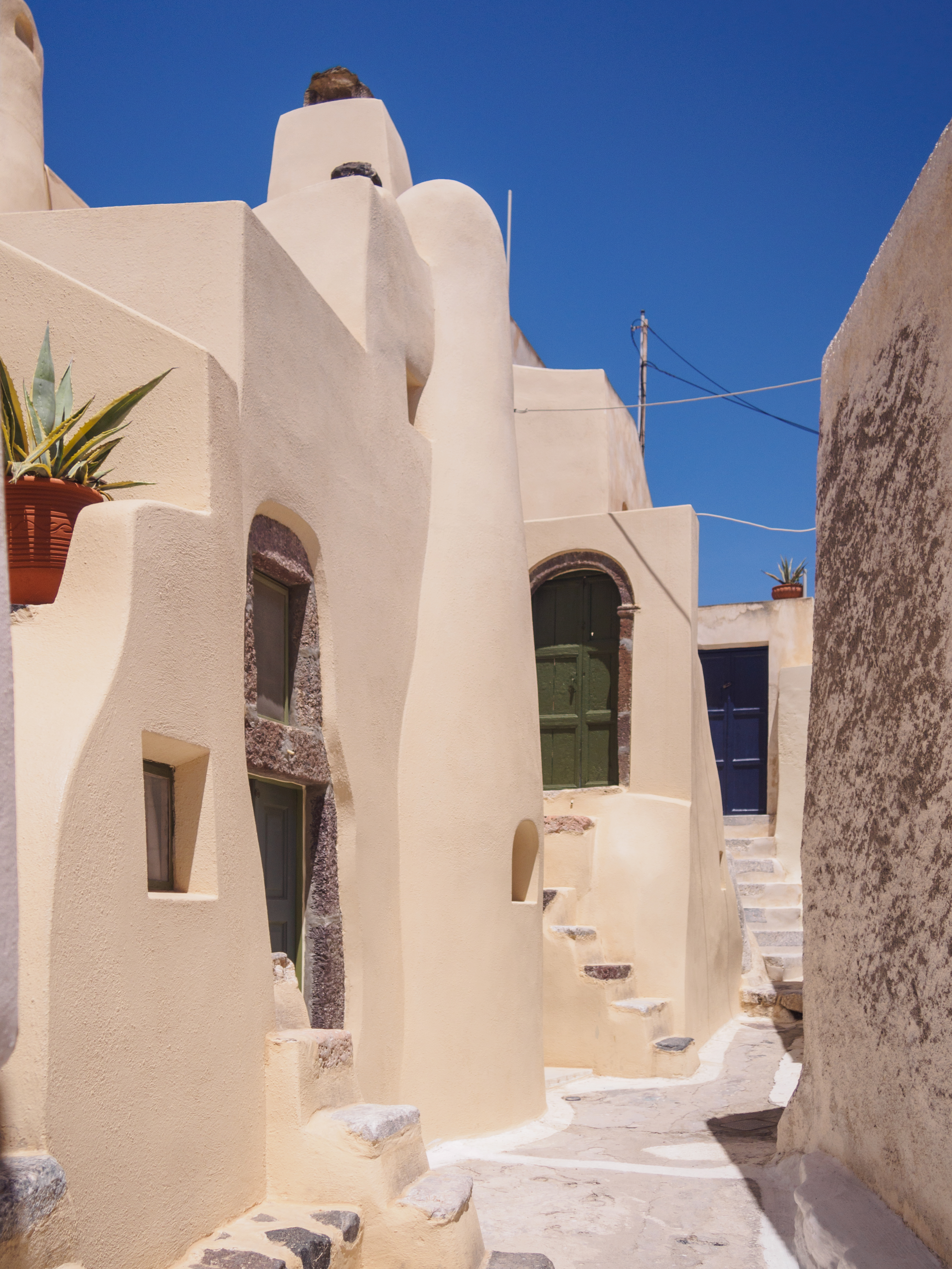 Une ruelle étroite d'un village cycladique aux murs crème arrondis et chaulés, une porte verte et une porte bleue, des marches de pierre, un agave en pot, sous un ciel bleu vif — le dédale médiéval d'Emporio (Kasteli), à Santorin.