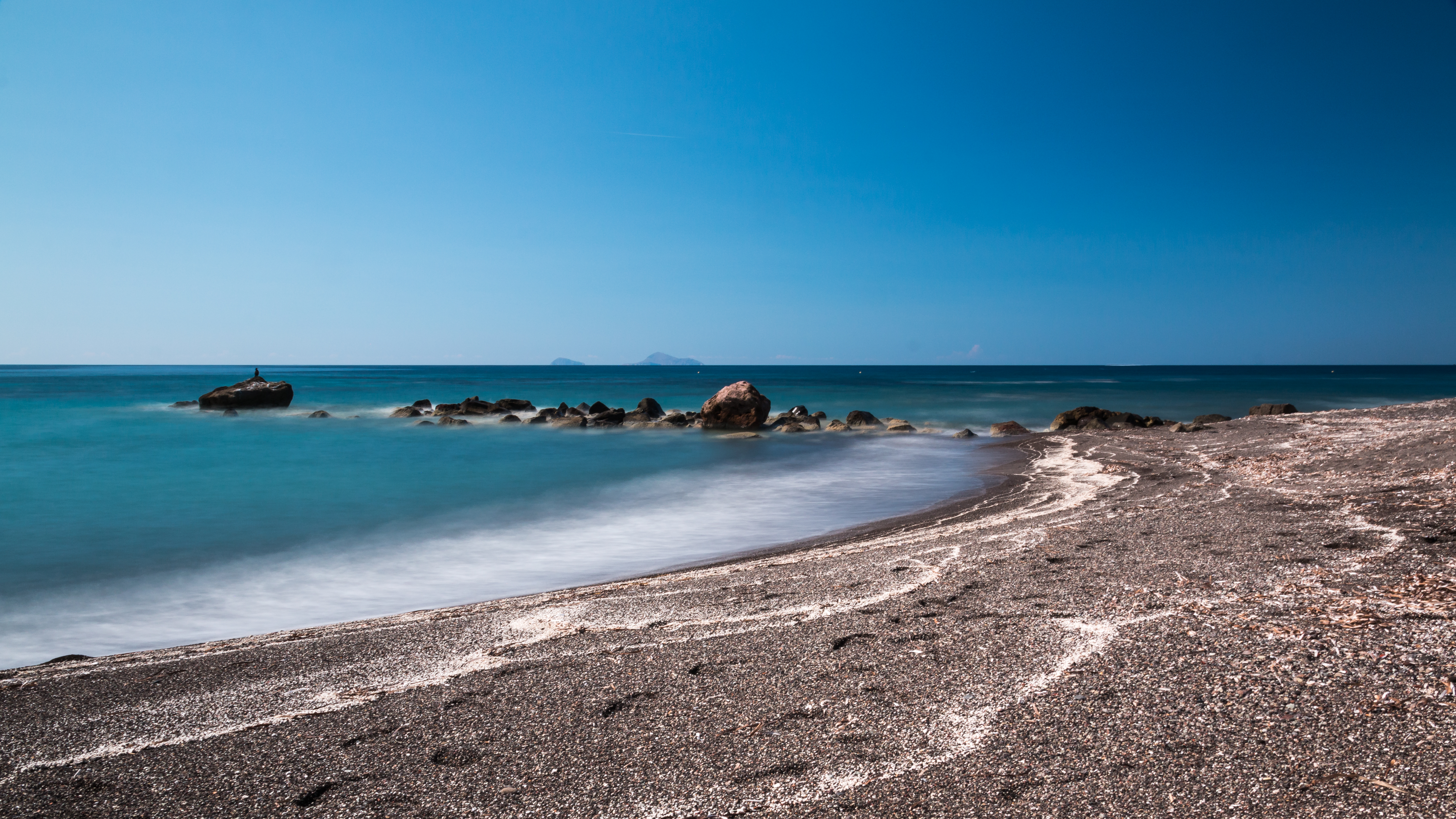 Une plage de sable et galets noirs volcaniques décrivant une longue courbe, une eau turquoise près du rivage virant au bleu profond, des rochers sombres affleurant dans l'eau, une petite île à l'horizon, sous un ciel bleu dégagé — la plage de Vlychada, au sud de Santorin.