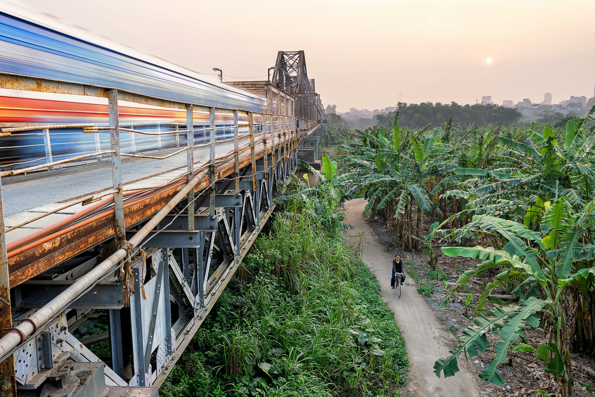 Pont Long Biên au coucher du soleil — train en flou de mouvement à gauche, cycliste sur chemin de terre parmi des bananiers à droite, lumière orangée et skyline de Hanoi en fond