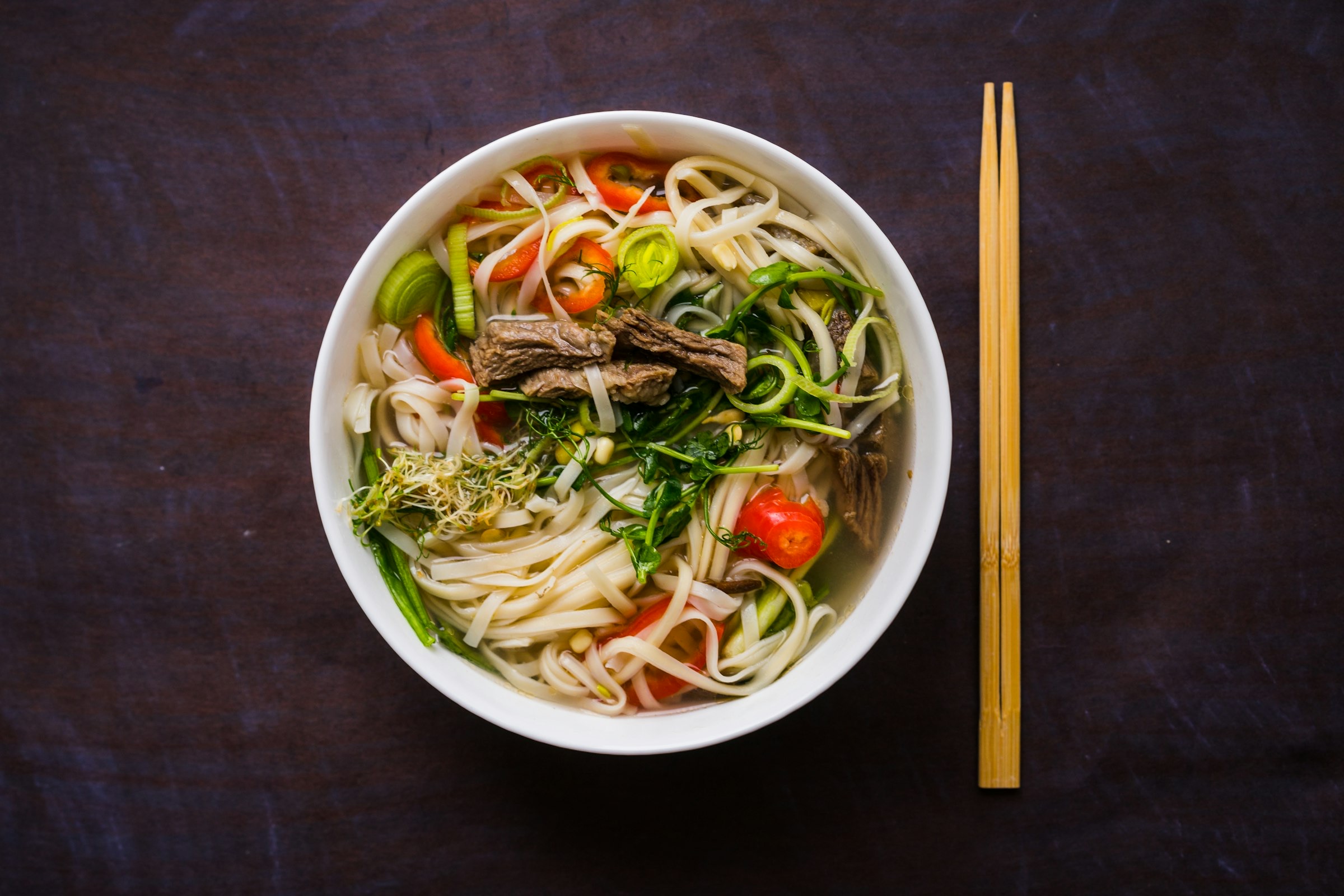 Bol de phở vu du dessus sur fond sombre — nouilles de riz blanches, tranches de bœuf, herbes fraîches, pousses de soja et bouillon clair, baguettes en bois à droite