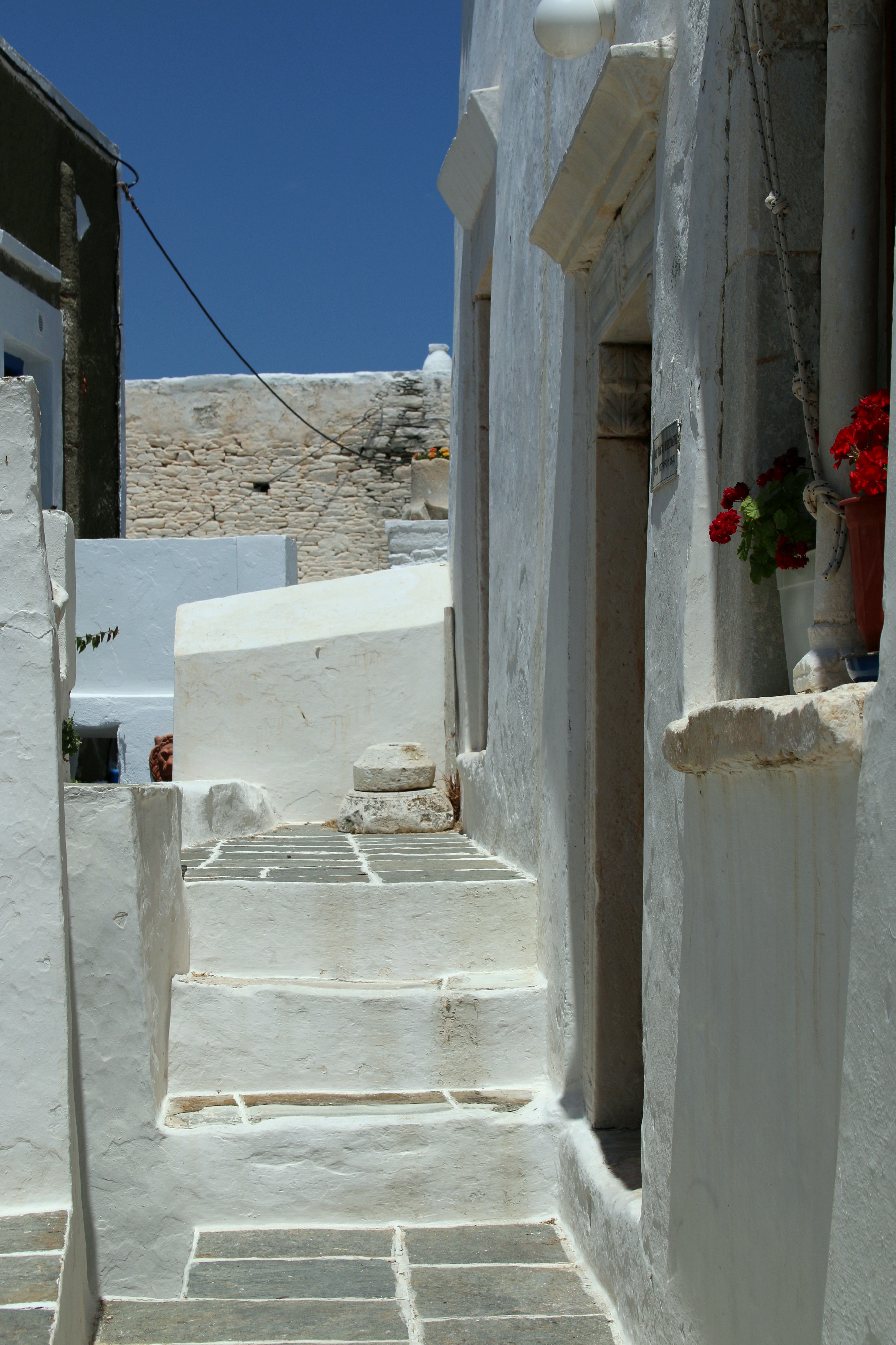 Une ruelle blanchie à la chaux du village médiéval de Kastro, à Sifnos : des marches de pierre peintes en blanc, une porte avec des géraniums rouges en pot, sous un ciel bleu