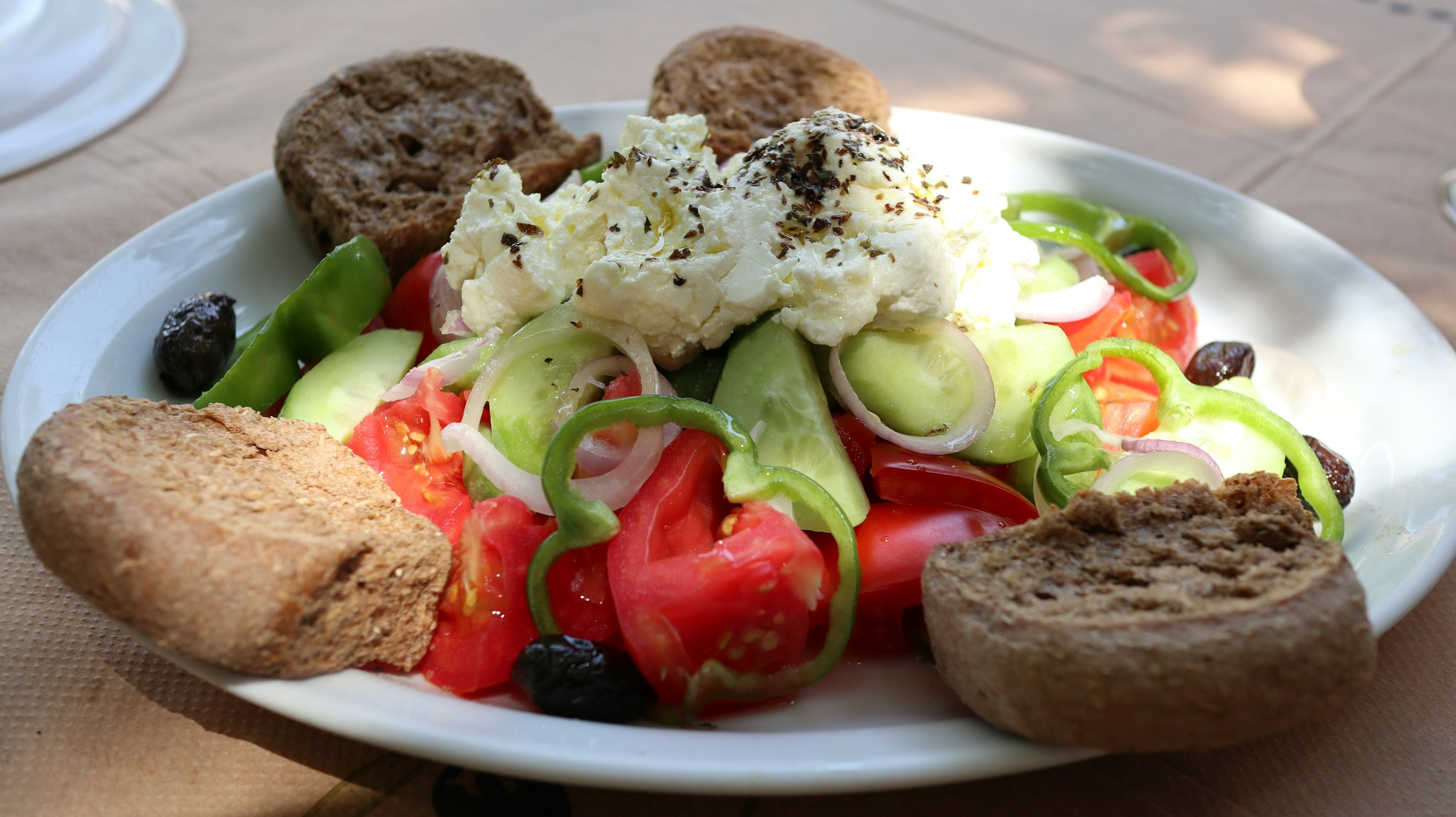 Une salade crétoise — tomates, concombre, poivron, oignon rouge, olives noires et myzithra à l'origan — entourée de paximadi, les biscottes d'orge, sur une assiette blanche.