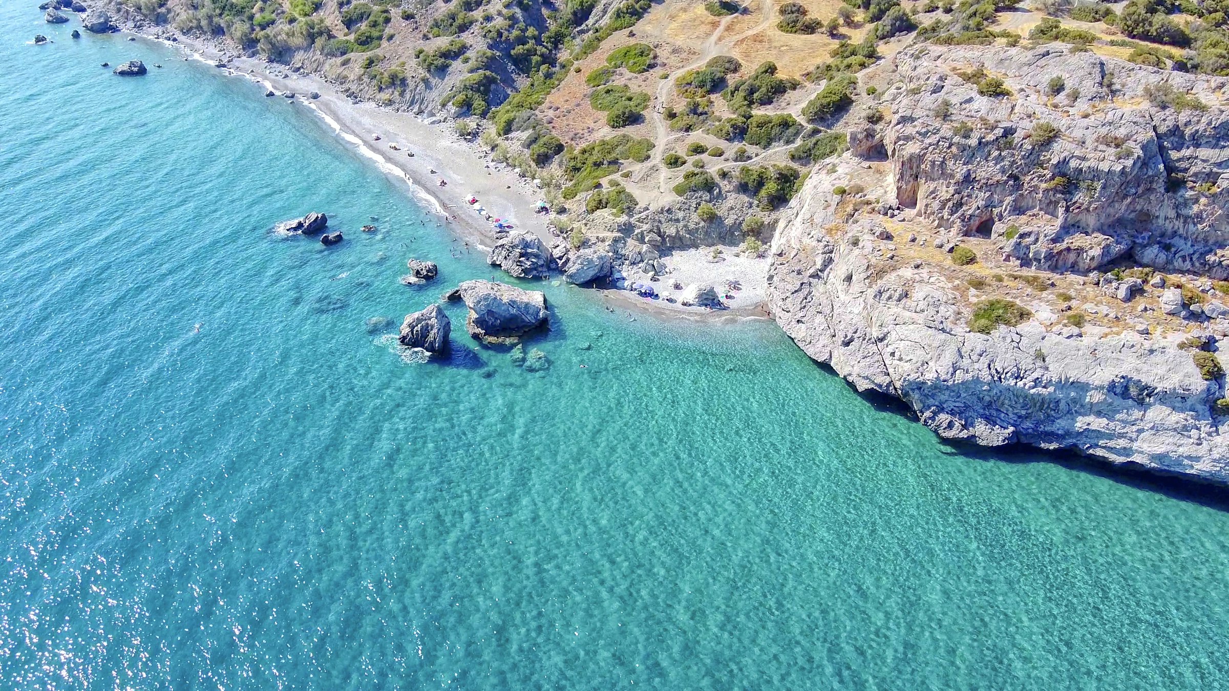 Vue aérienne d'une plage de galets et de rochers au pied de collines arides, une eau turquoise cristalline — côte sud de la Crète.